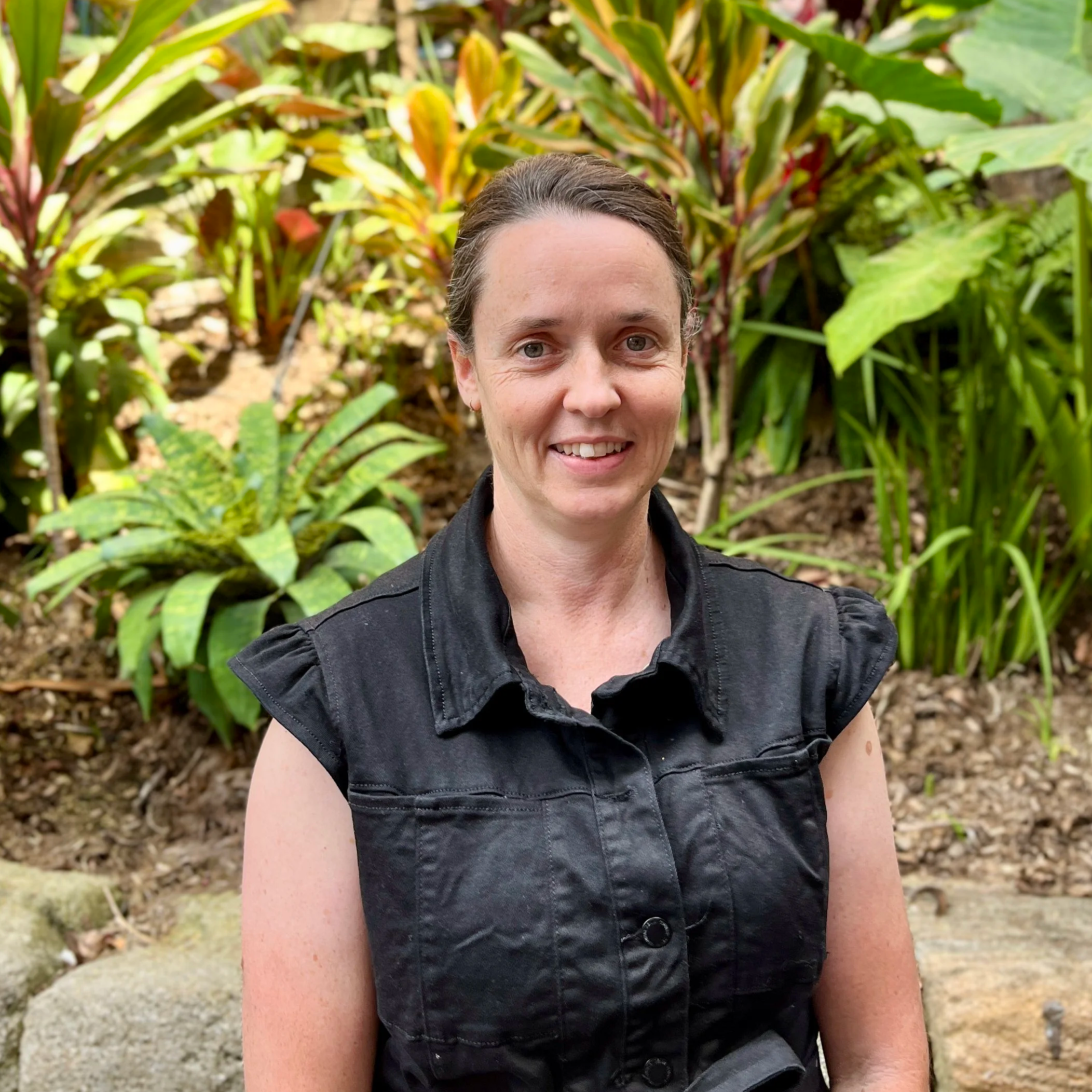 A woman with dark hair, fair skin, and freckles, smiling, wearing a sleeveless black floral top, standing against a plain off-white wall.