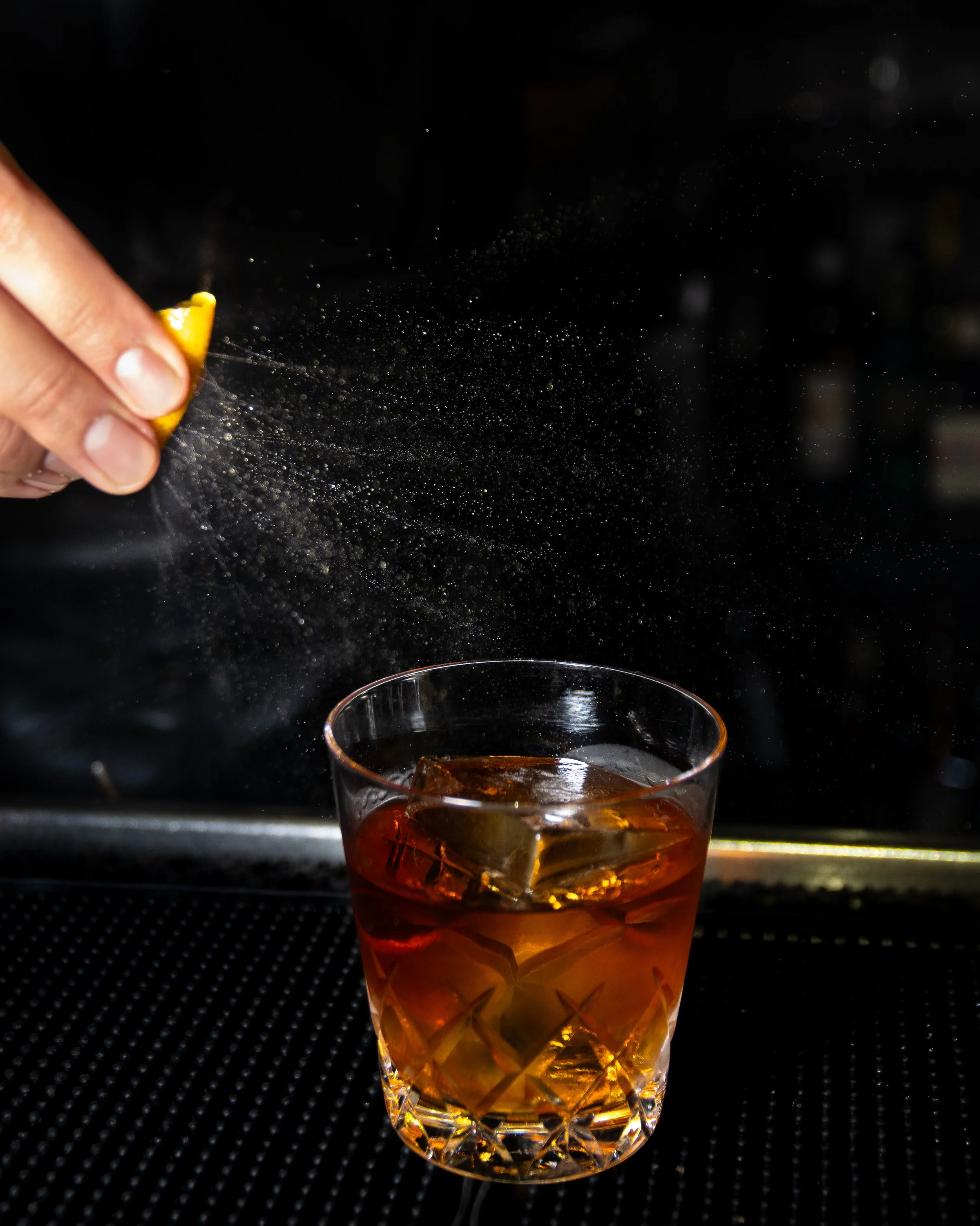 A person sprinkles a lemon peel into a glass of whiskey with ice, against a dark background.
