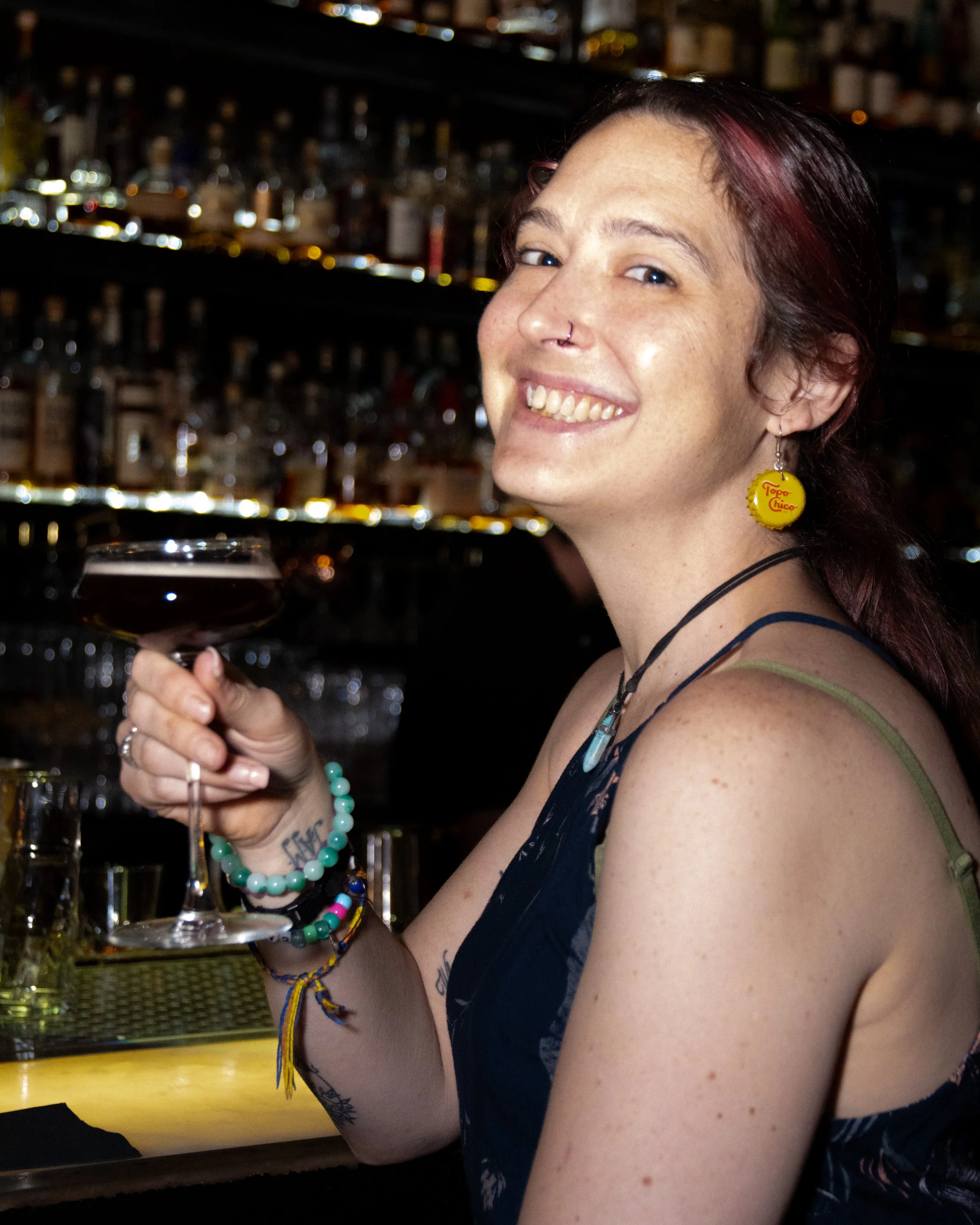 A smiling woman with red hair and a nose ring holding a cocktail glass in a bar setting.