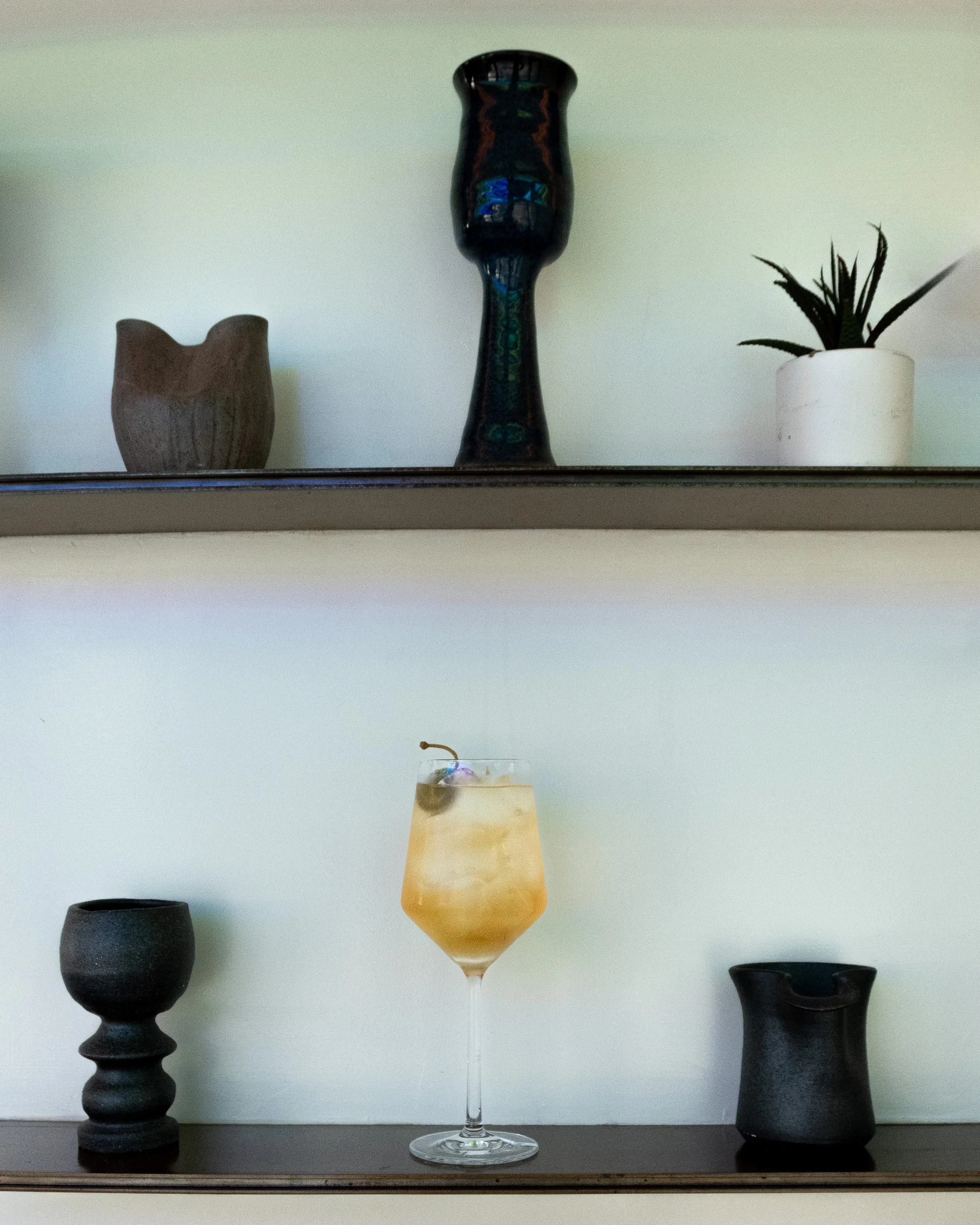 Shelf with a black cup, a yellow cocktail with ice and a caper berry on top, and a small black cup or vase beneath two shelves, with decorative objects including a wooden bowl, a tall dark vase with a face design, and a potted plant.