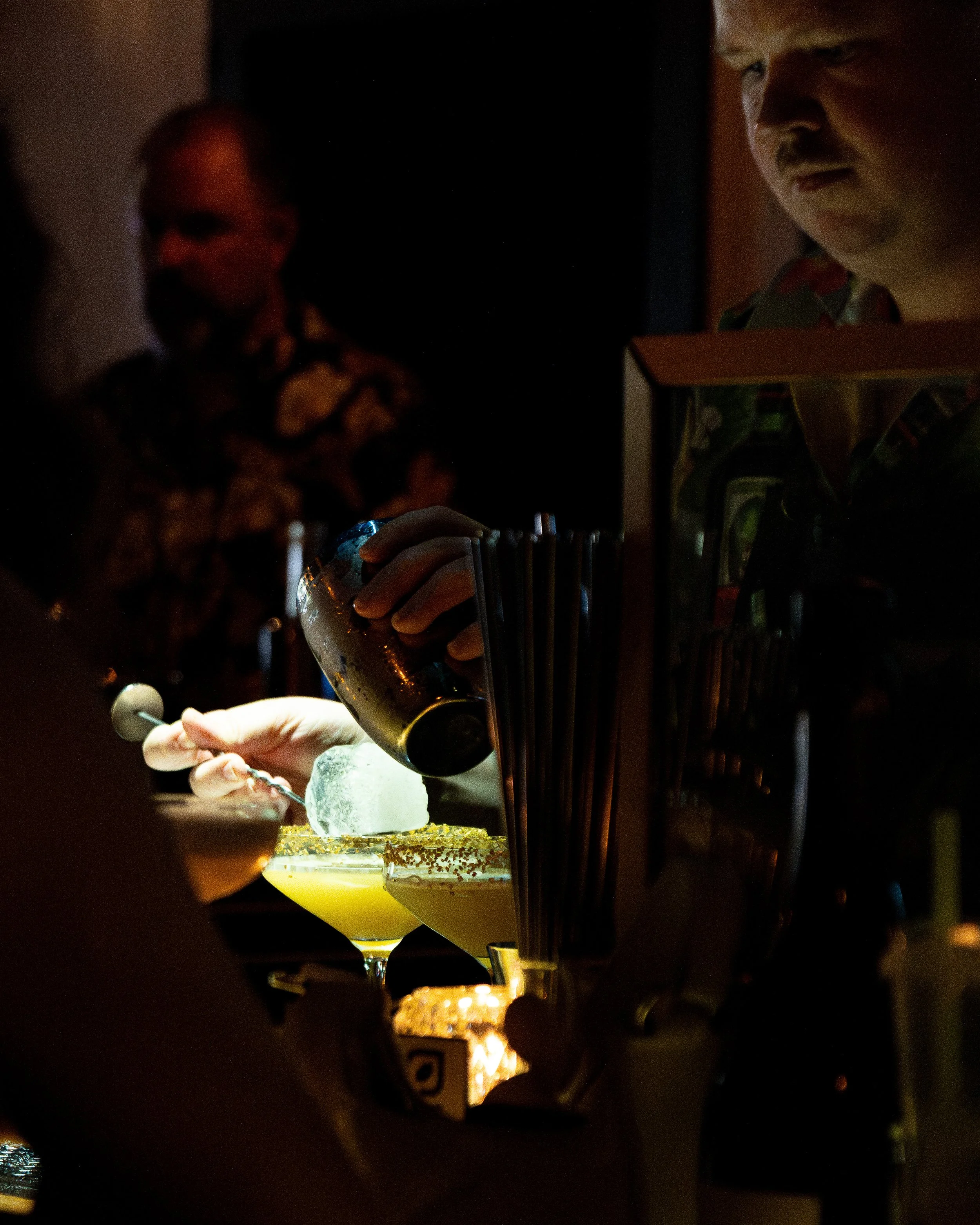 A bartender pouring a drink into a cocktail glass, with patrons in the background in a dimly lit bar.