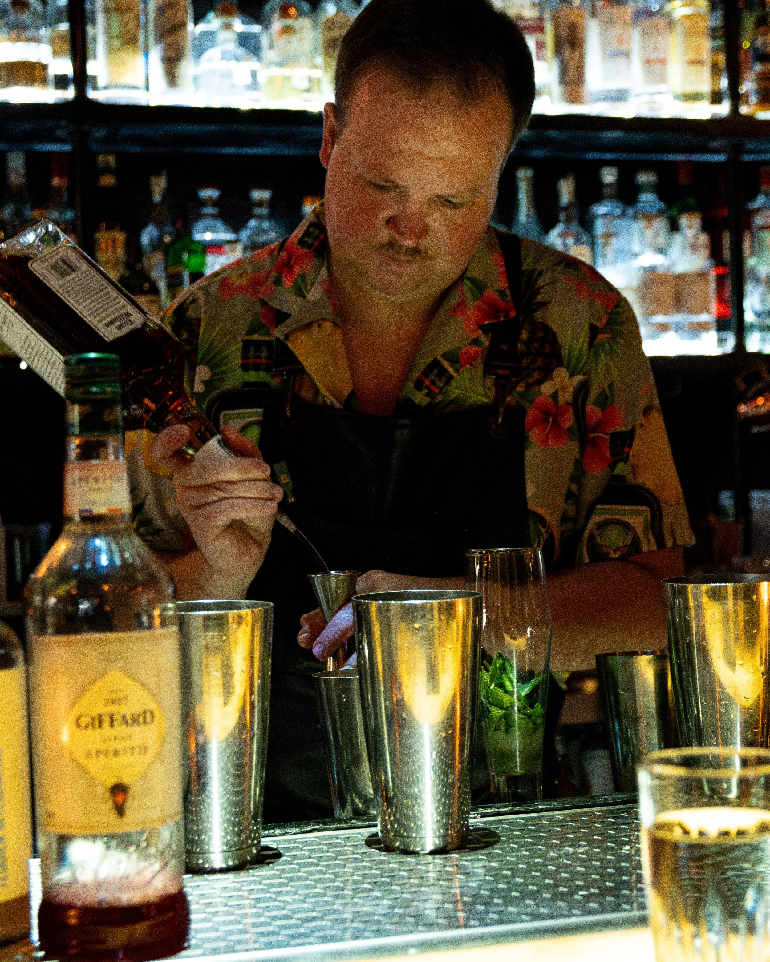 A bartender with a mustache wearing a colorful Hawaiian shirt and black apron pours a drink into a metal shaker at a bar, surrounded by various bottles and glasses.
