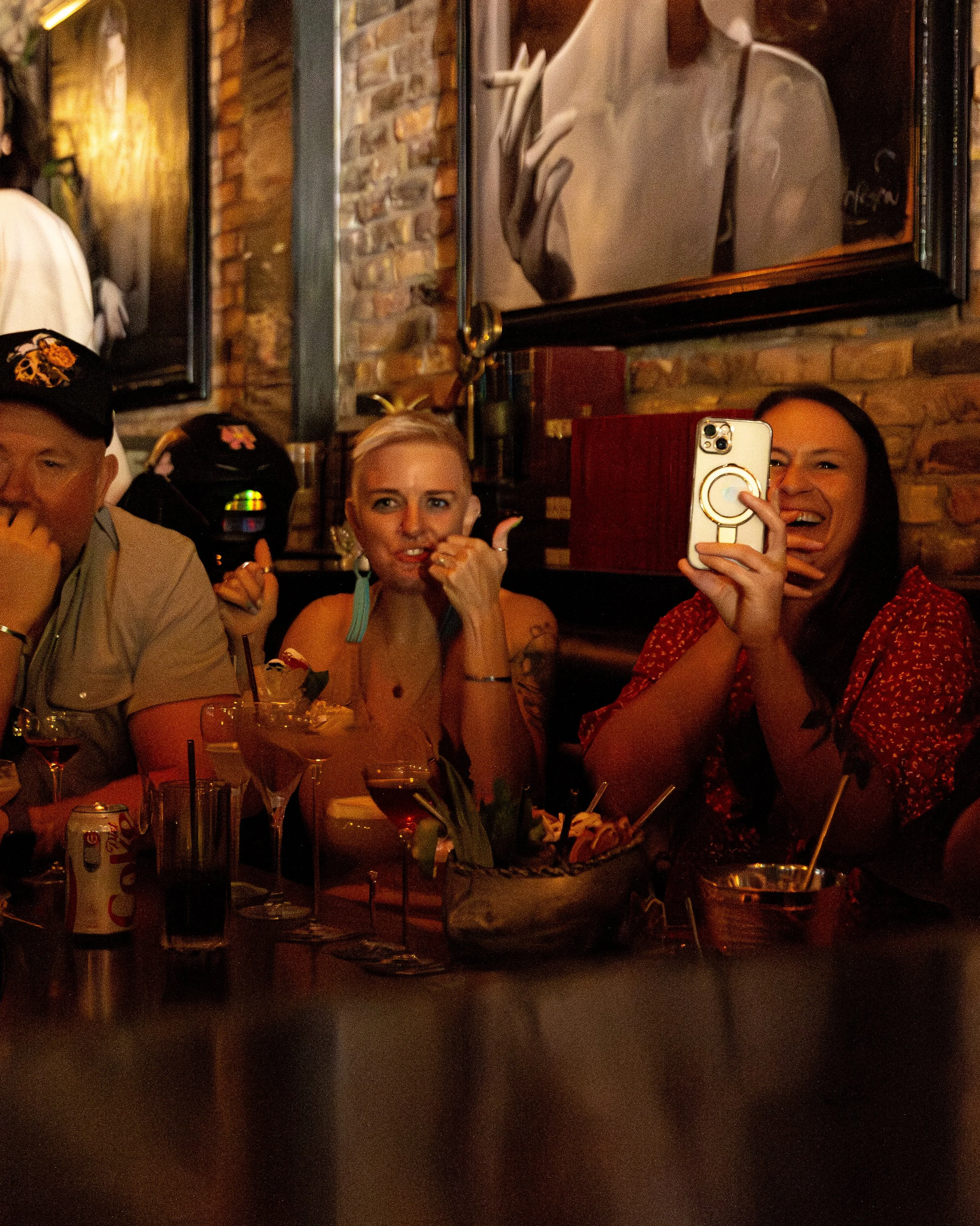 Group of people sitting at a table in a dimly lit bar or restaurant, taking photos and smiling, with drinks and snacks on the table, and framed black-and-white photographs on the brick wall behind them.