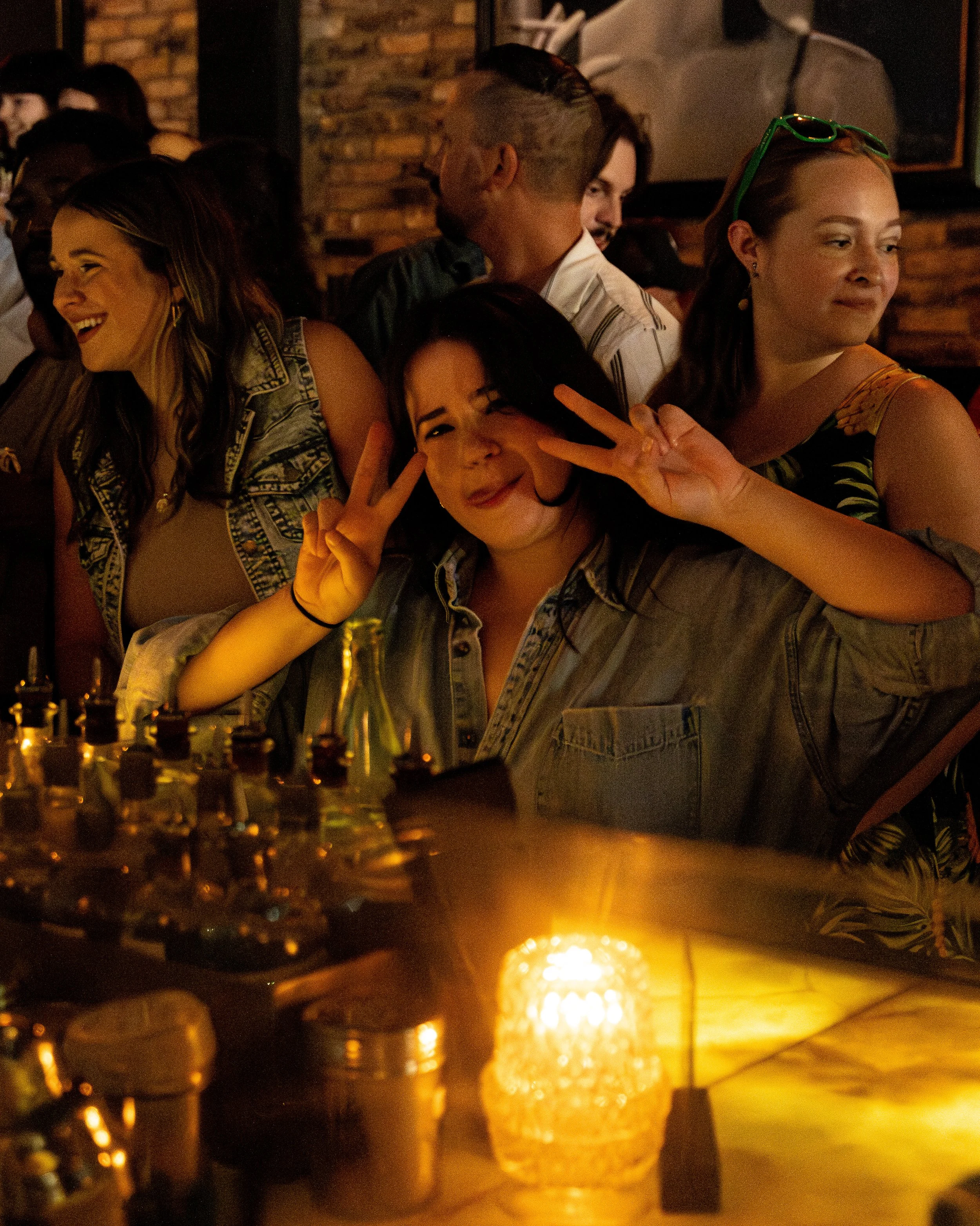 Group of young women at a bar, one making a peace sign and smiling at the camera, others talking and laughing, warm ambient lighting, background brick wall.
