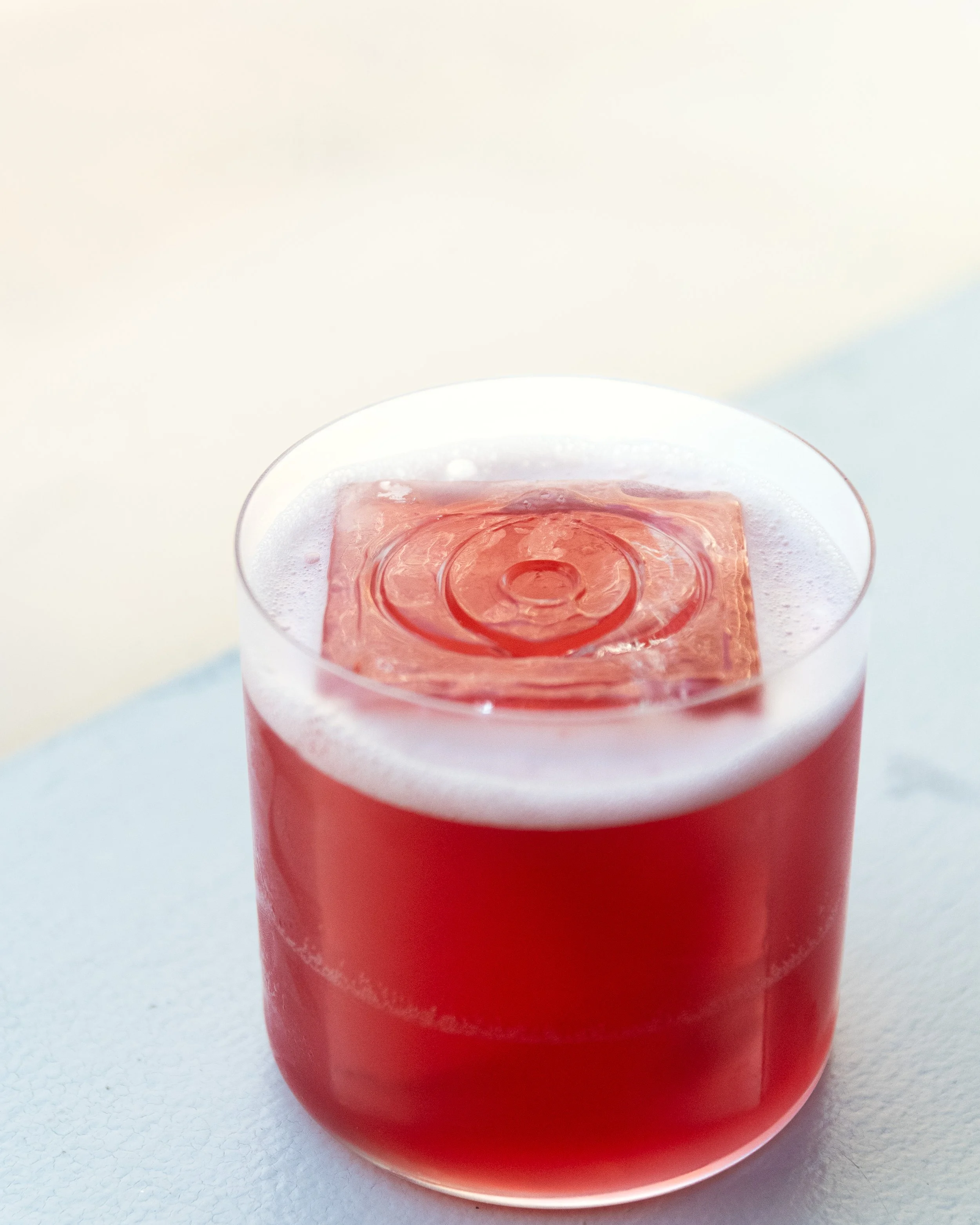 A clear glass filled with red-colored liquid and a large ice cube on top.
