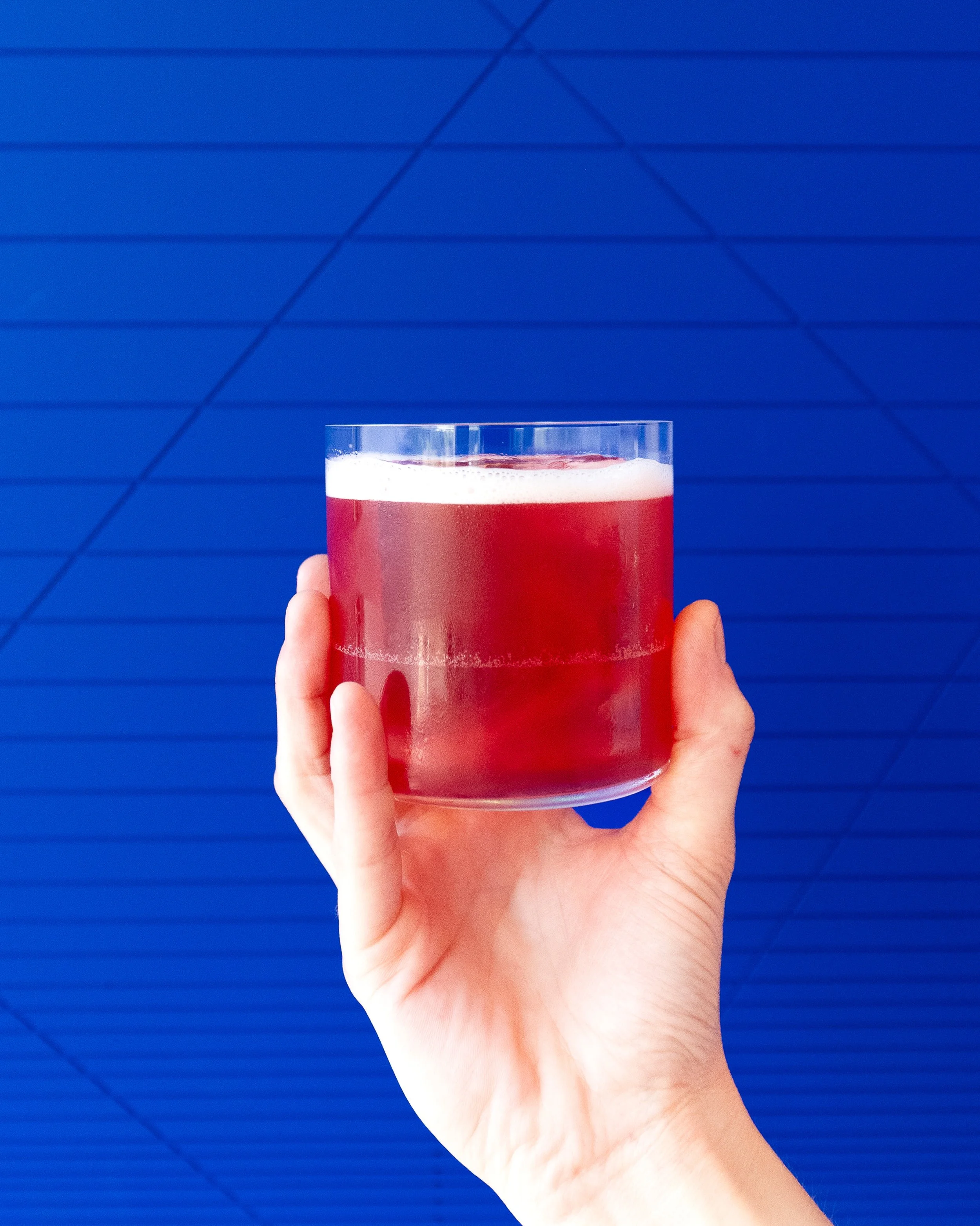 A hand holding a transparent glass with a red beverage against a blue tiled background.