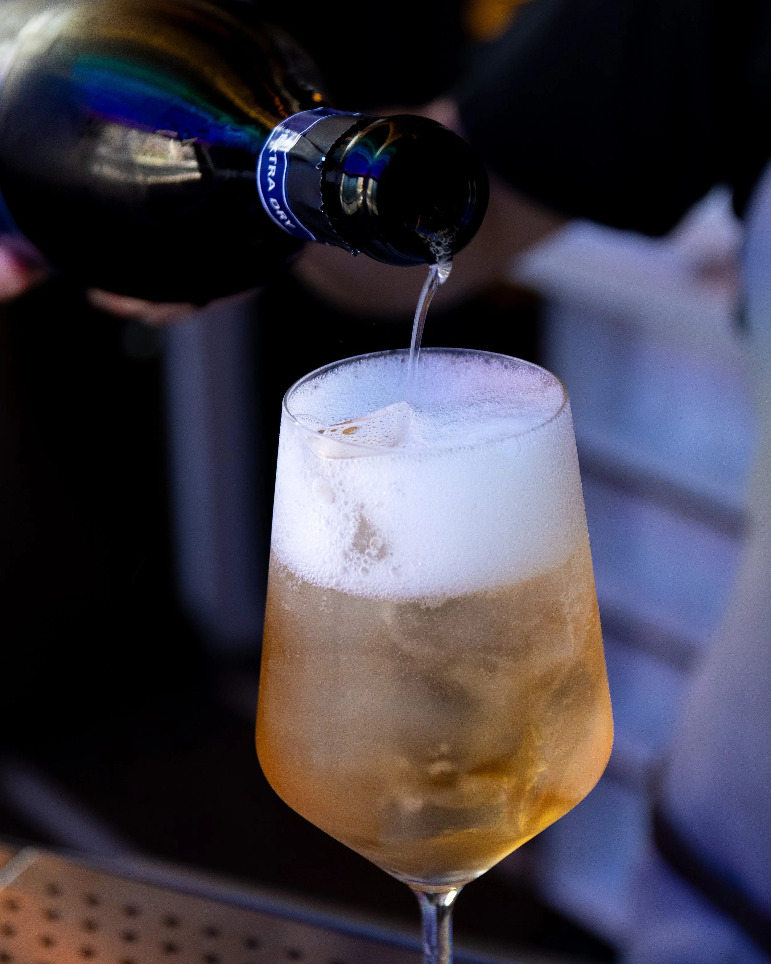 A person pouring champagne into a tall glass with ice, creating foam on top.