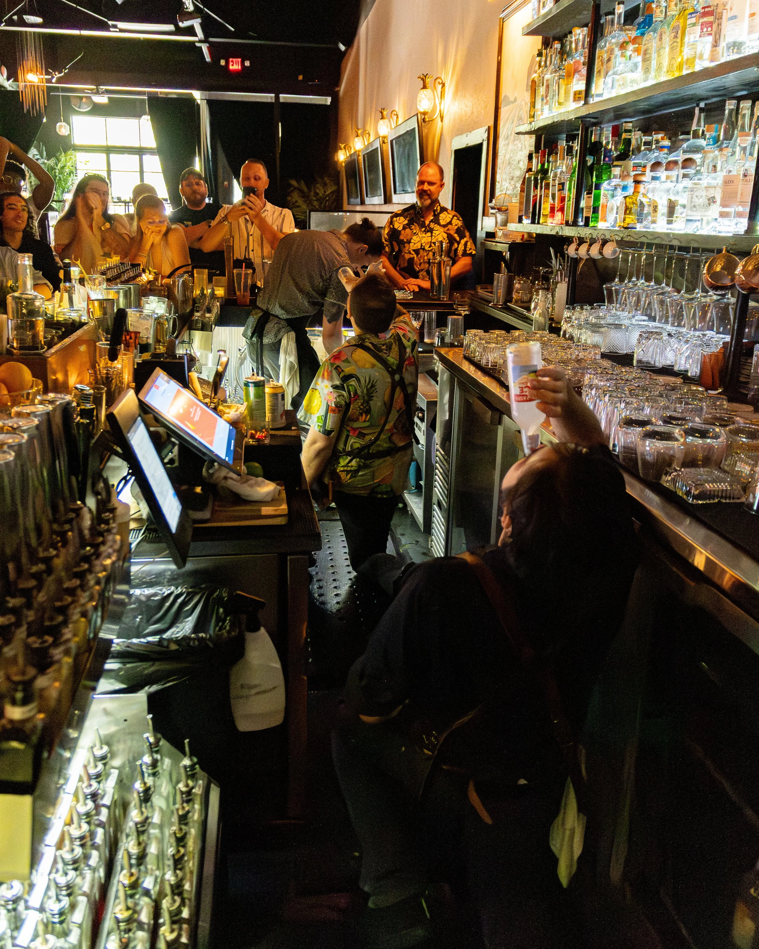 Crowded bar scene with bartenders preparing drinks, bar shelves stocked with alcohol bottles, and patrons watching the staff. Dim lighting and a lively atmosphere.