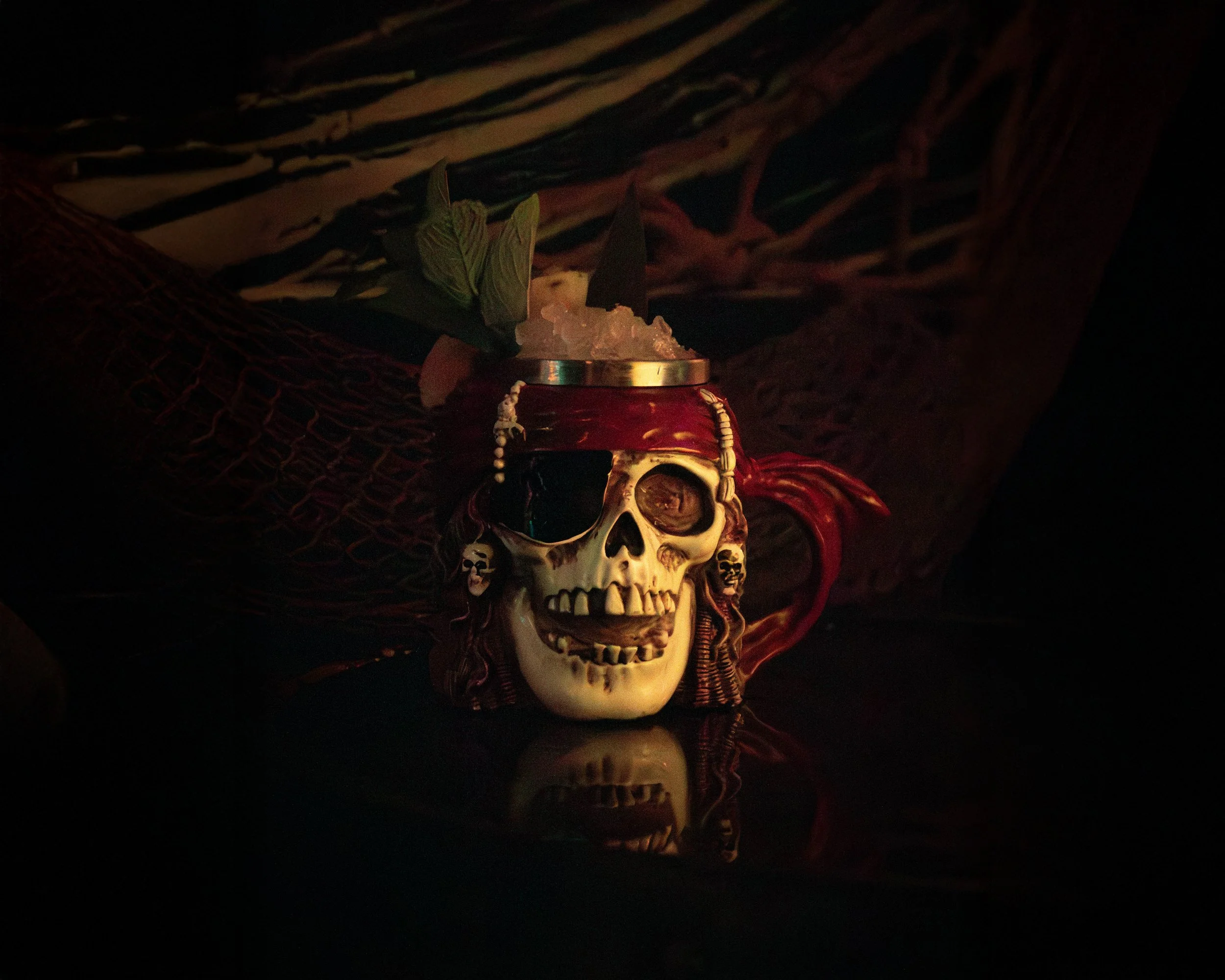 Decorative skull mug with a straw, flowers, and attached pirate earrings, illuminated in a dim setting, reflecting on a dark surface.