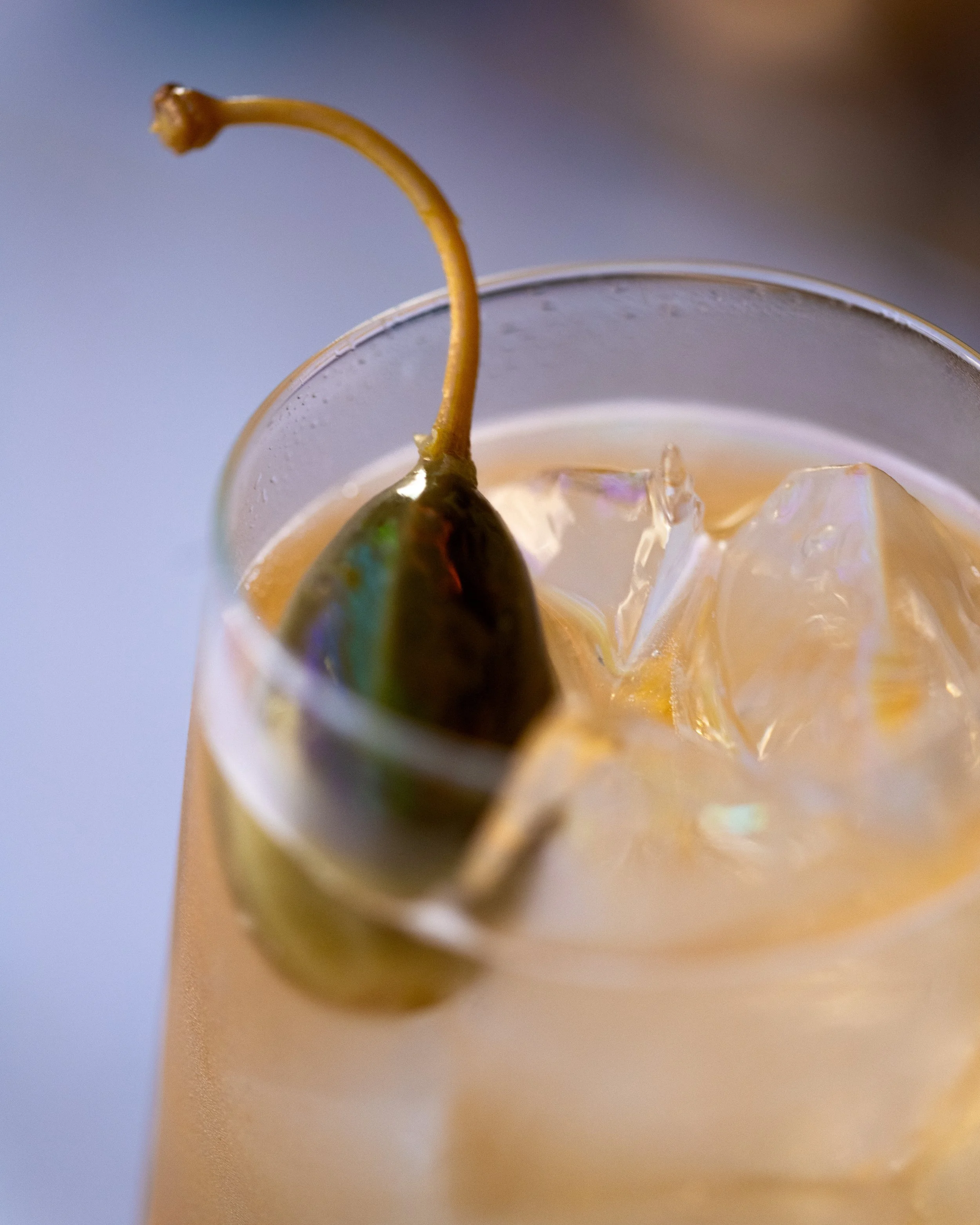Close-up of a cocktail with ice, a lime wedge, and a caper berry on the rim of the glass.