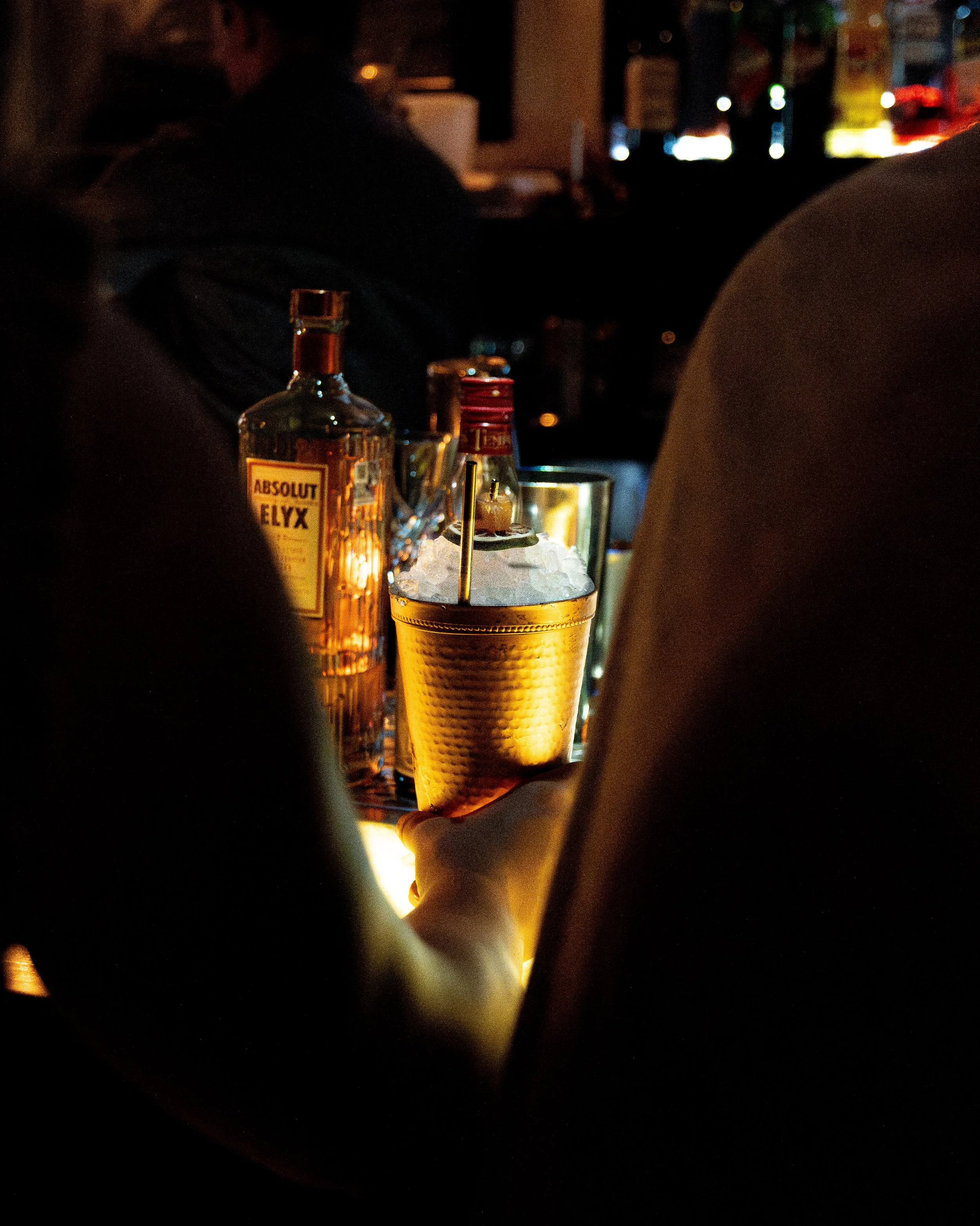 A bar scene with a focus on a cocktail glass with ice, a bottle of Absolut Elyx vodka, and a person's hand holding the drink. The background is dark with some colorful, blurry lights.
