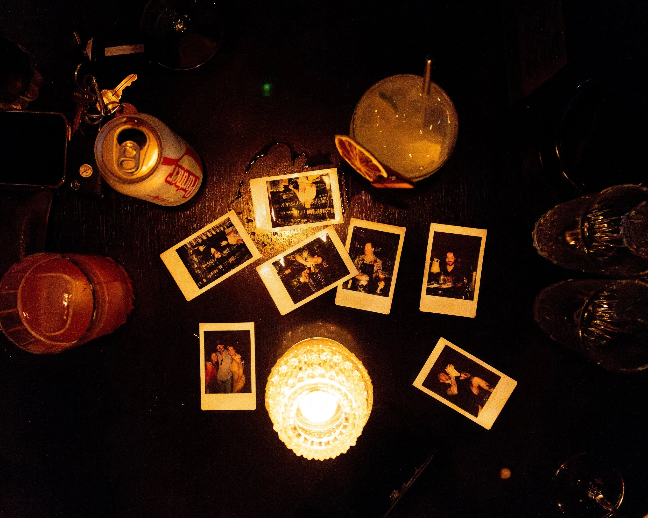 A dark table with a lit candle, scattered instant photo prints, a can of soda, two glasses of drinks, and a few additional glasses, all seen from above.