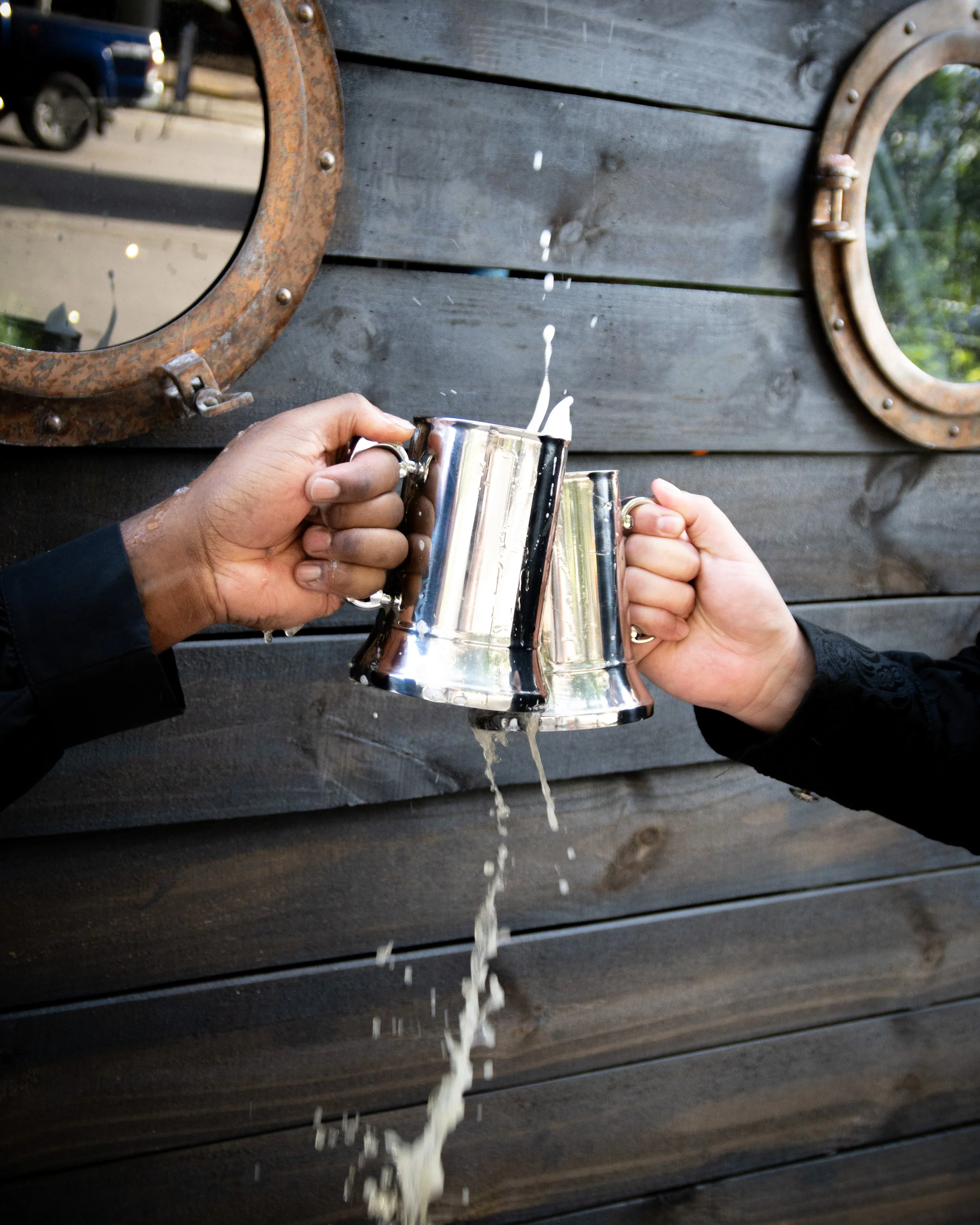 Two people clinking metallic mugs filled with a liquid, spilling over onto a dark wooden wall behind them.