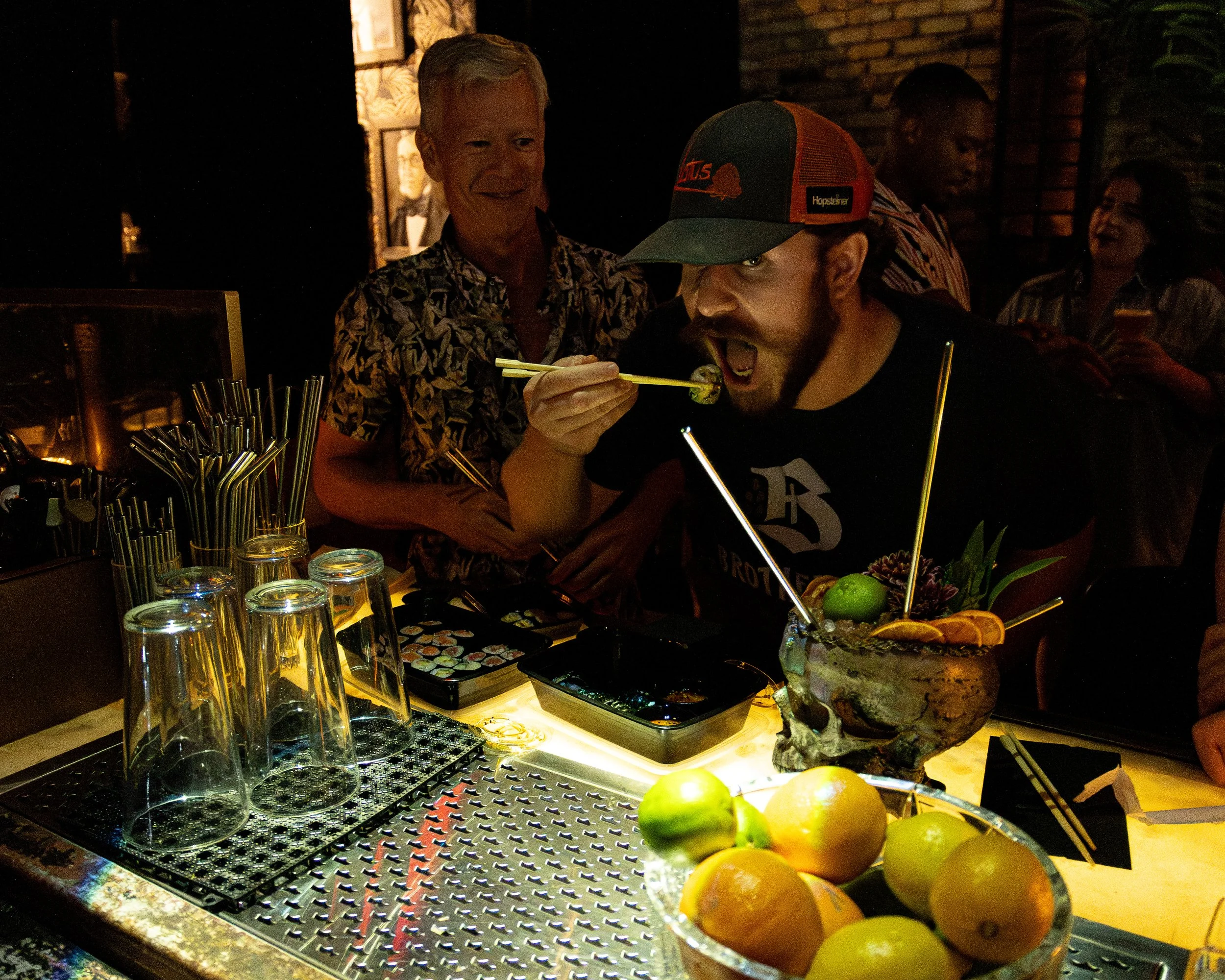 Man wearing a baseball cap eating sushi with chopsticks at a dimly lit bar, with a bowl of fresh fruit and drinks on the counter, and other people in the background.