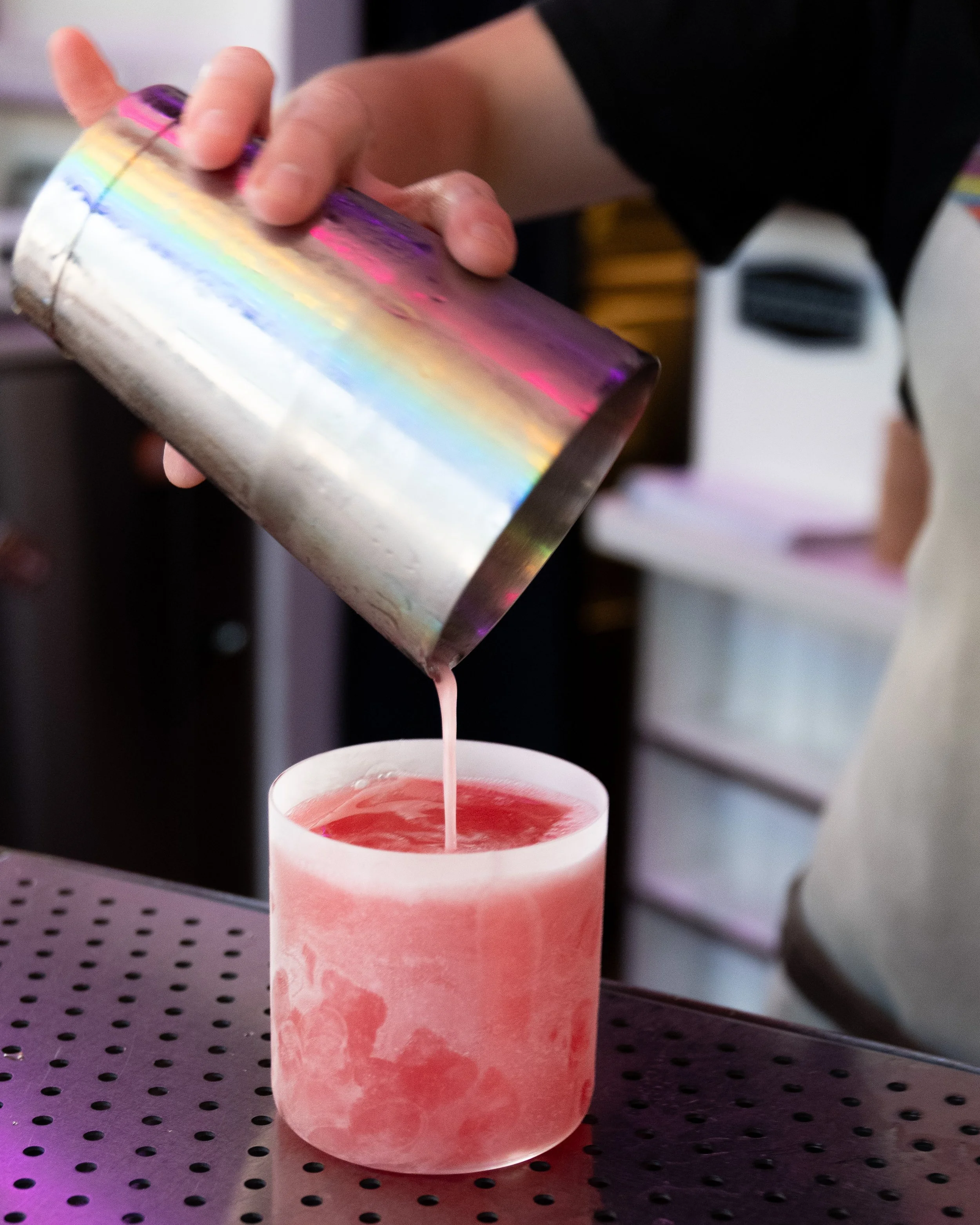 A person is pouring a pink beverage from a metallic rainbow-colored container into a pink frosted glass on a black perforated surface.