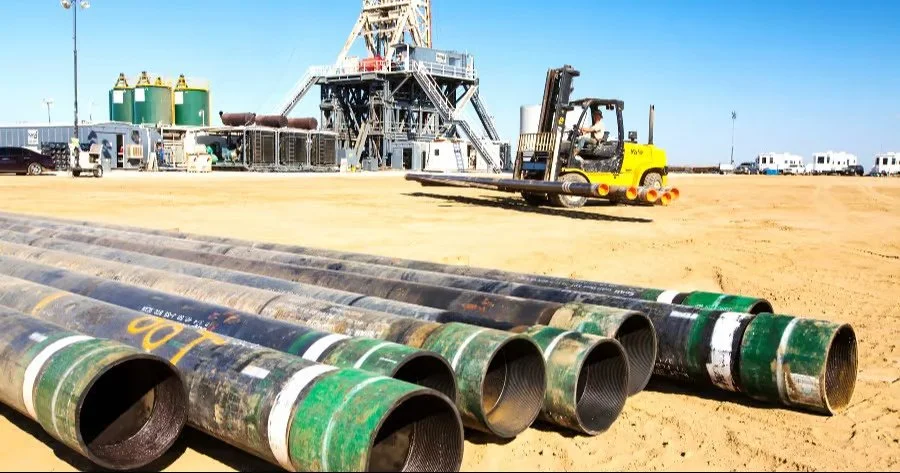 Oil drilling site with pipes in the foreground and a drilling rig in the background, along with a roller compactor on sandy ground.