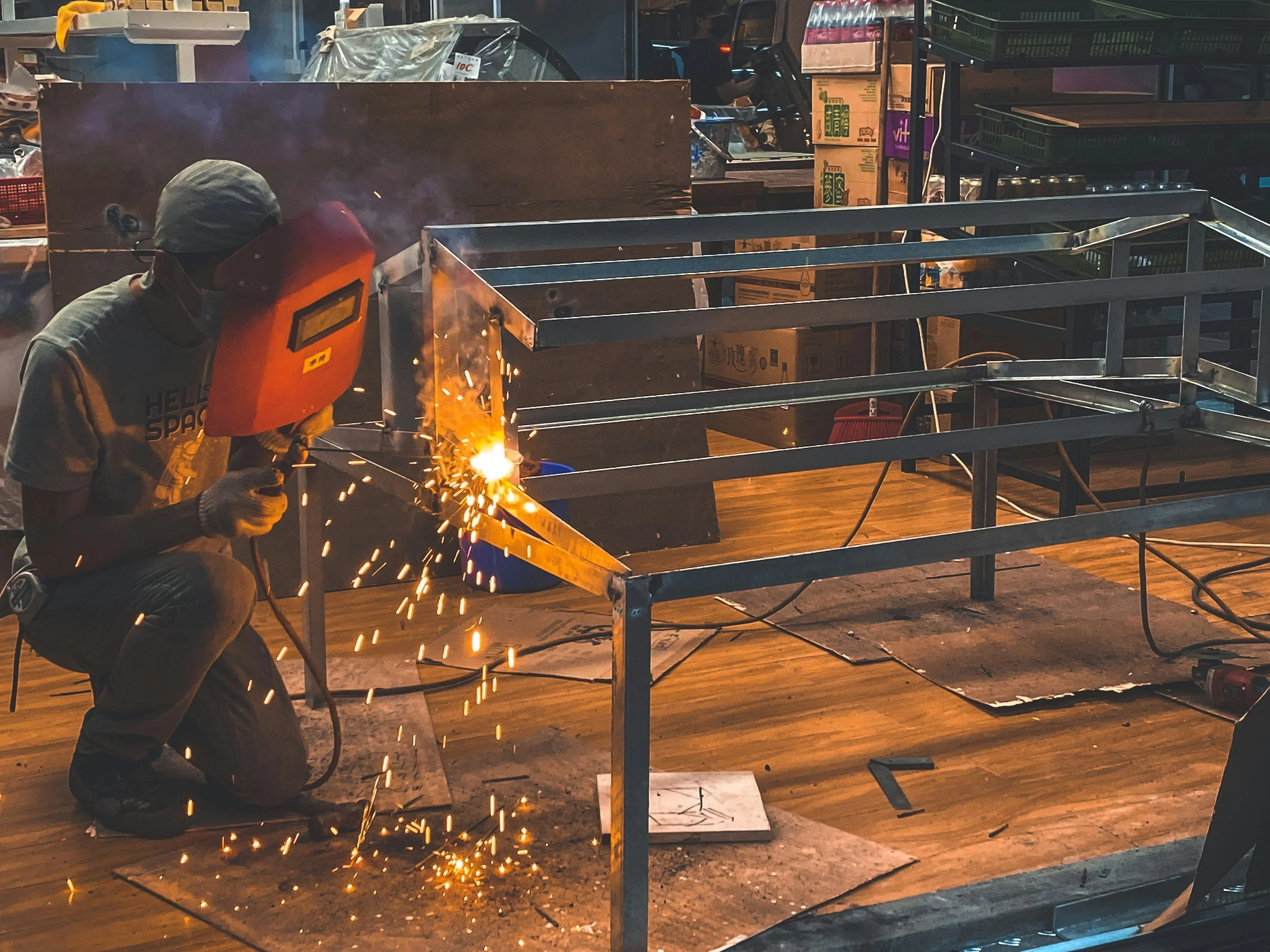 A person welding a metal frame in a workshop, sparks flying from the welding process, with shelves and boxes in the background.