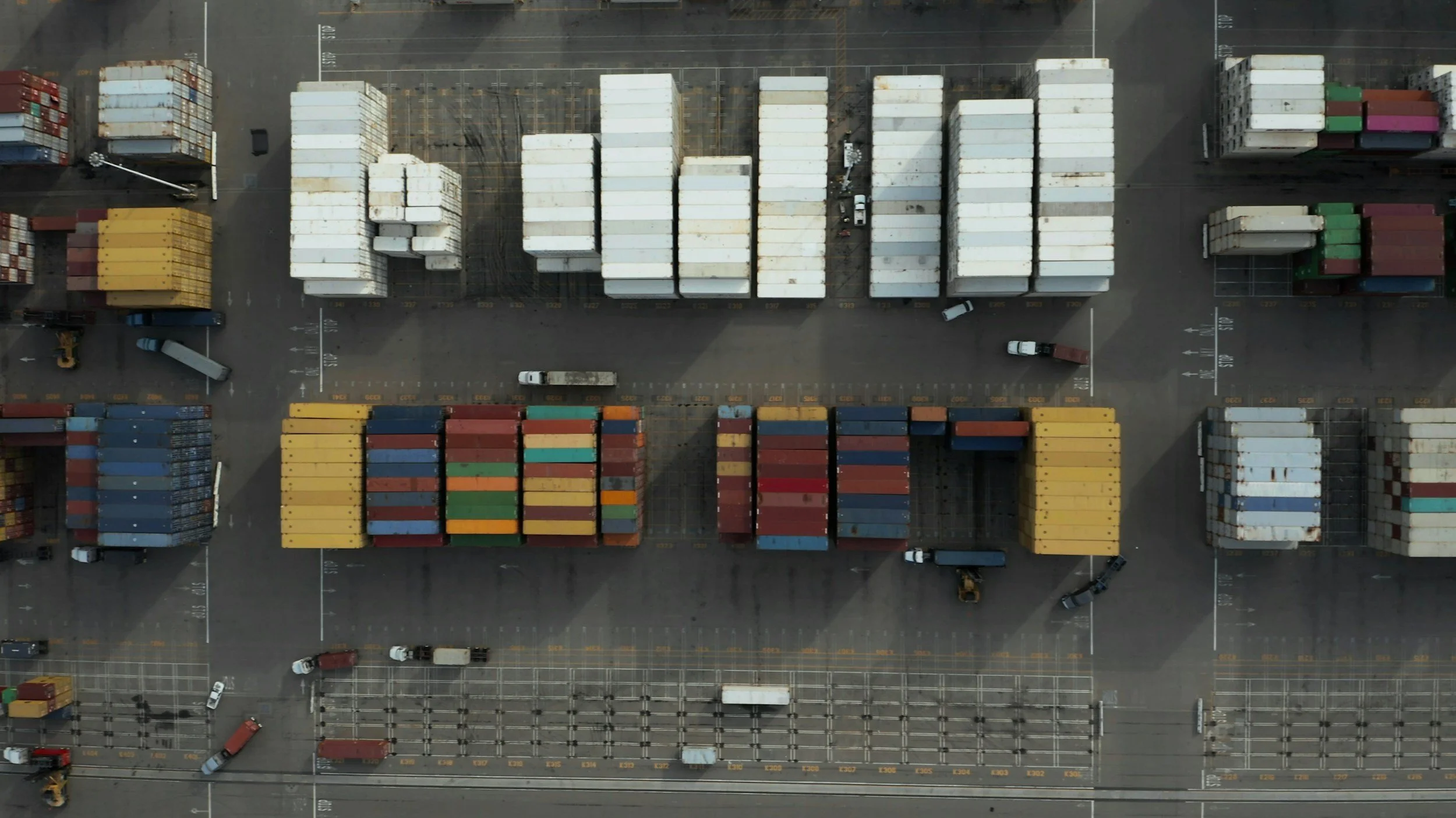 Bird's-eye view of a shipping container yard with containers stacked in rows and trucks parked among the containers.