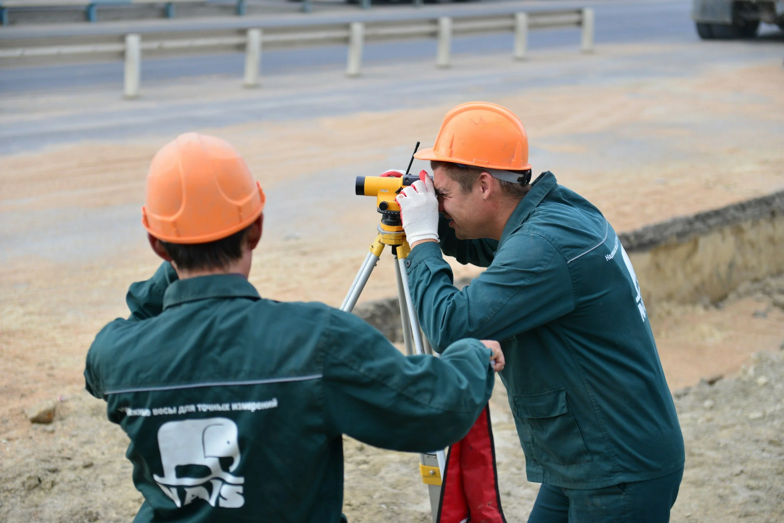 Two construction workers wearing orange safety helmets and green uniforms using a surveyor's tools at a construction site near a road.