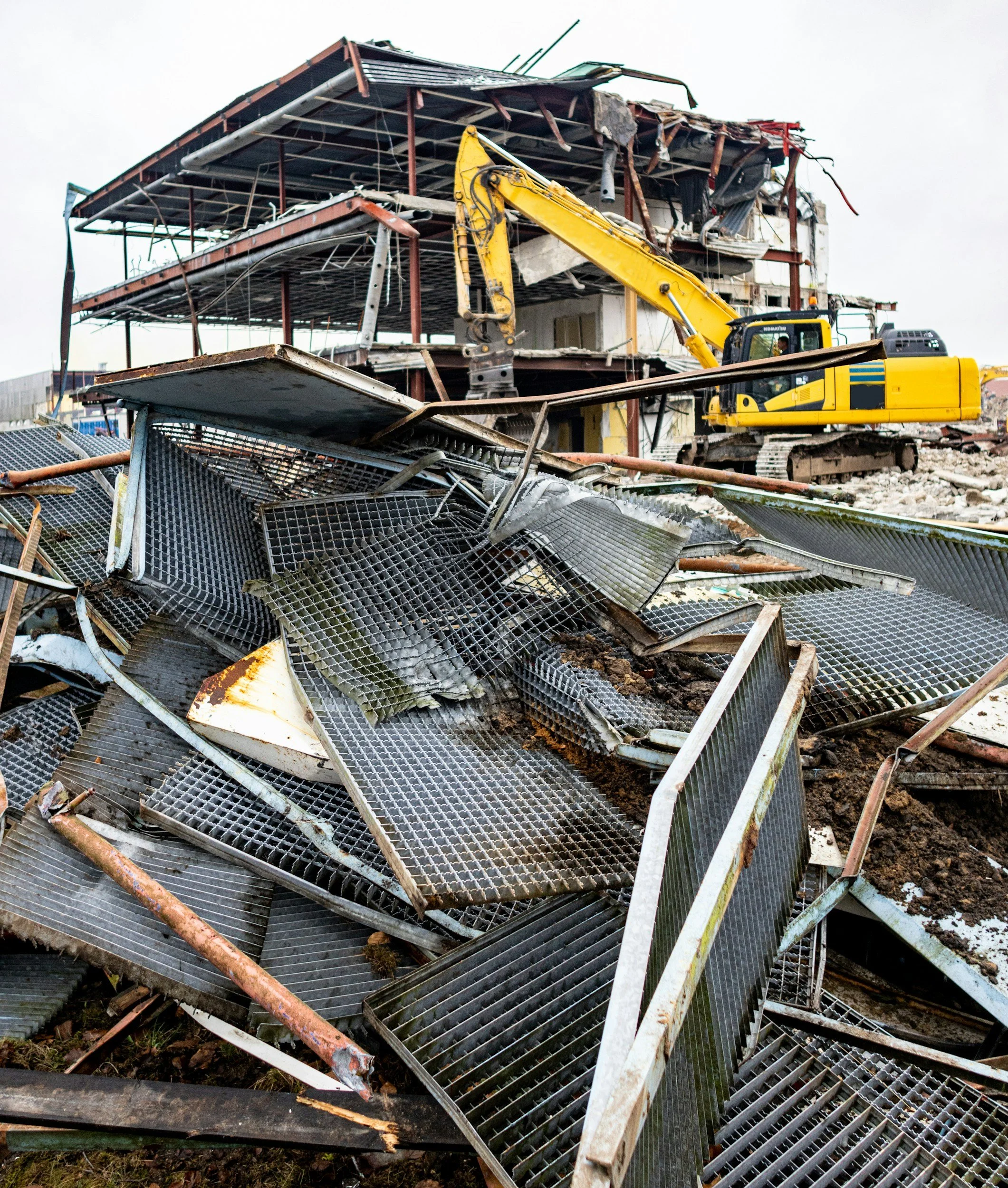 Debris from a destroyed building with metal framework and a yellow excavator amidst rubble.