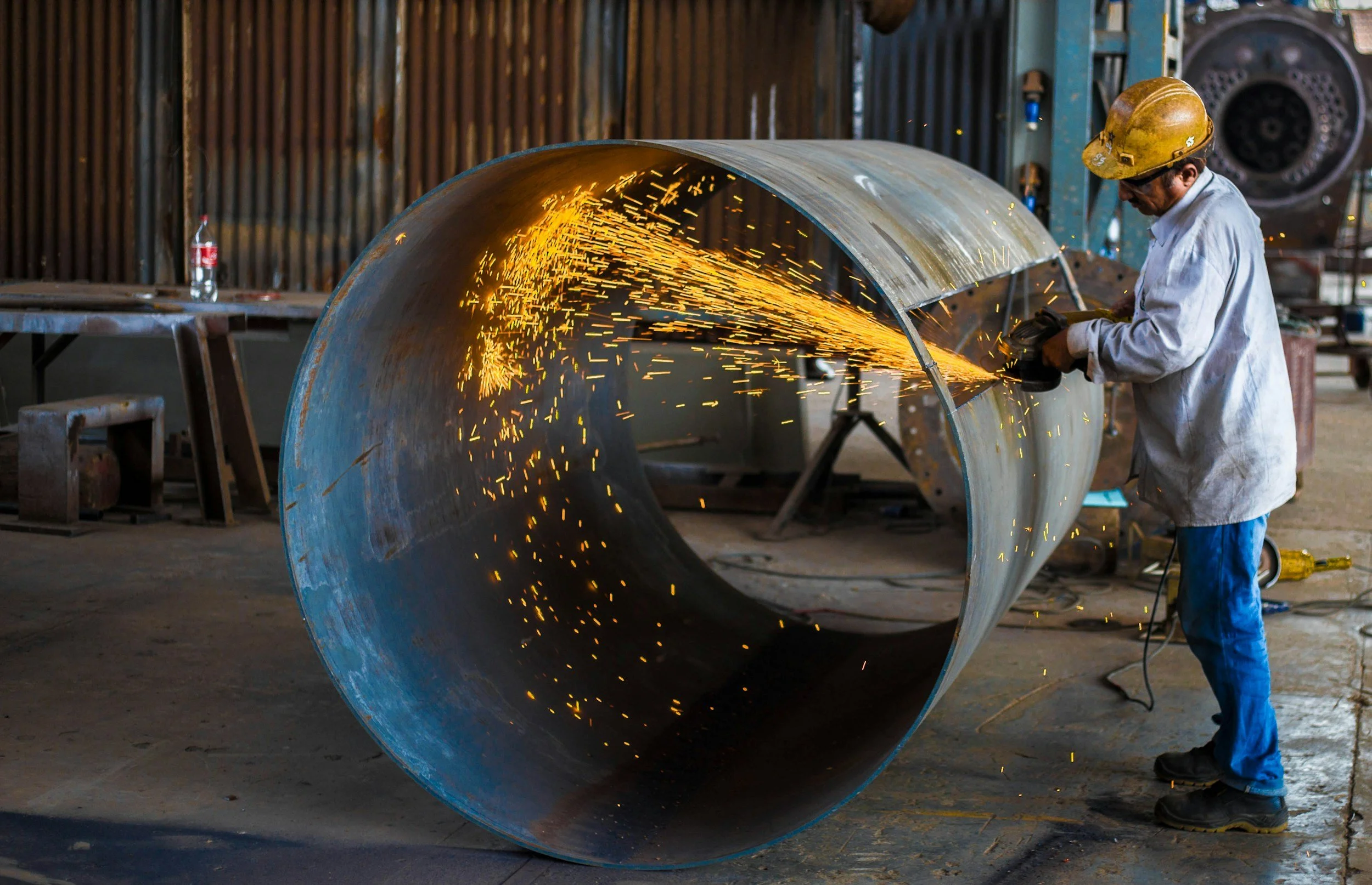 A worker wearing a yellow hard hat, white safety coat, and blue jeans is welding a large metal pipe inside a factory or workshop. Sparks are flying from the welding process.