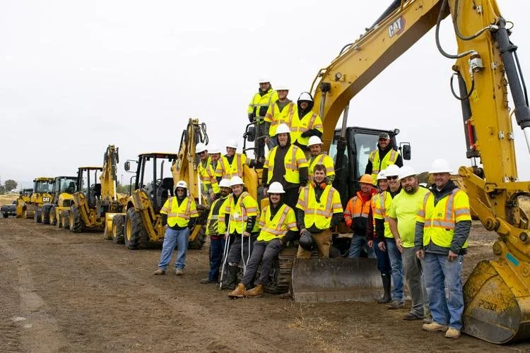 Group of construction workers in yellow safety vests and hard hats standing on and around a row of yellow heavy machinery on a construction site.