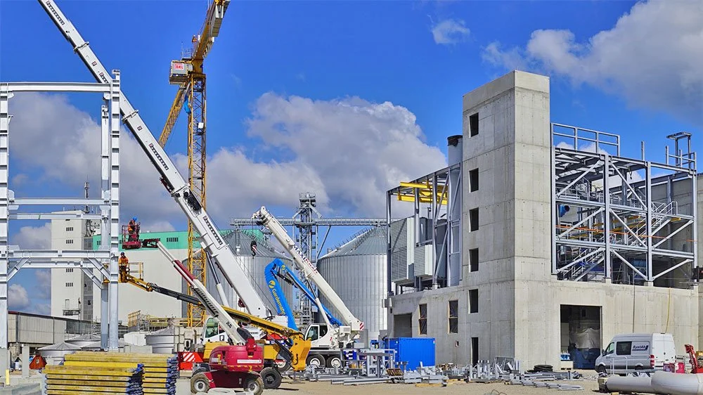 Construction site with cranes, scaffolding, and unfinished buildings under a blue sky.