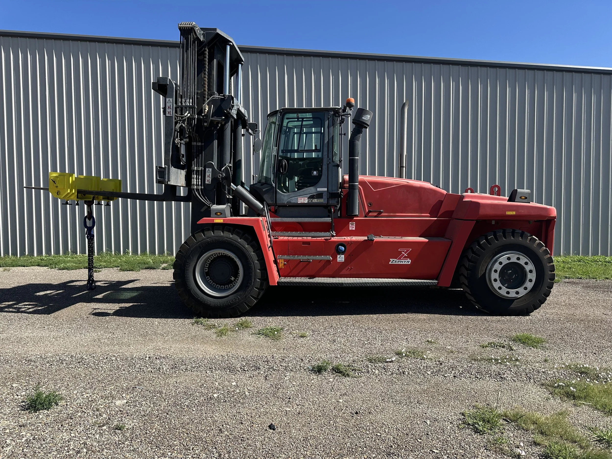 A large red forklift parked on a gravel surface in front of a metal building under a clear blue sky.