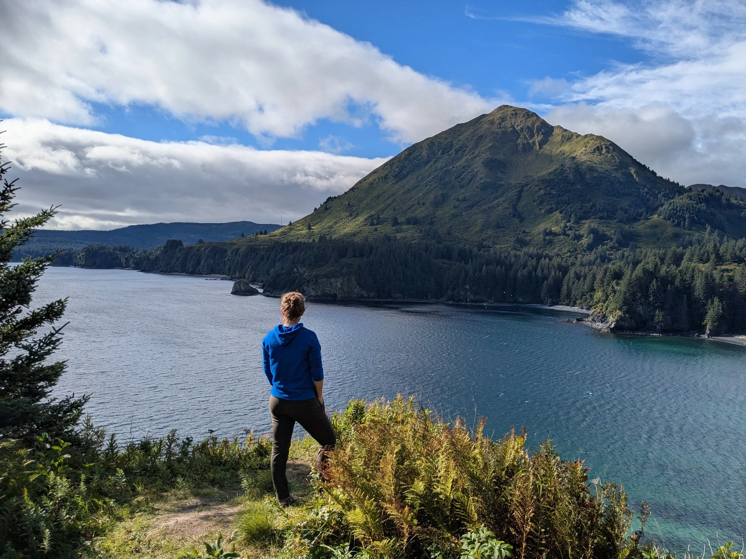 A person dressed in a blue jacket and dark pants standing near a body of water, overlooking a lush green mountain under a partly cloudy sky.
