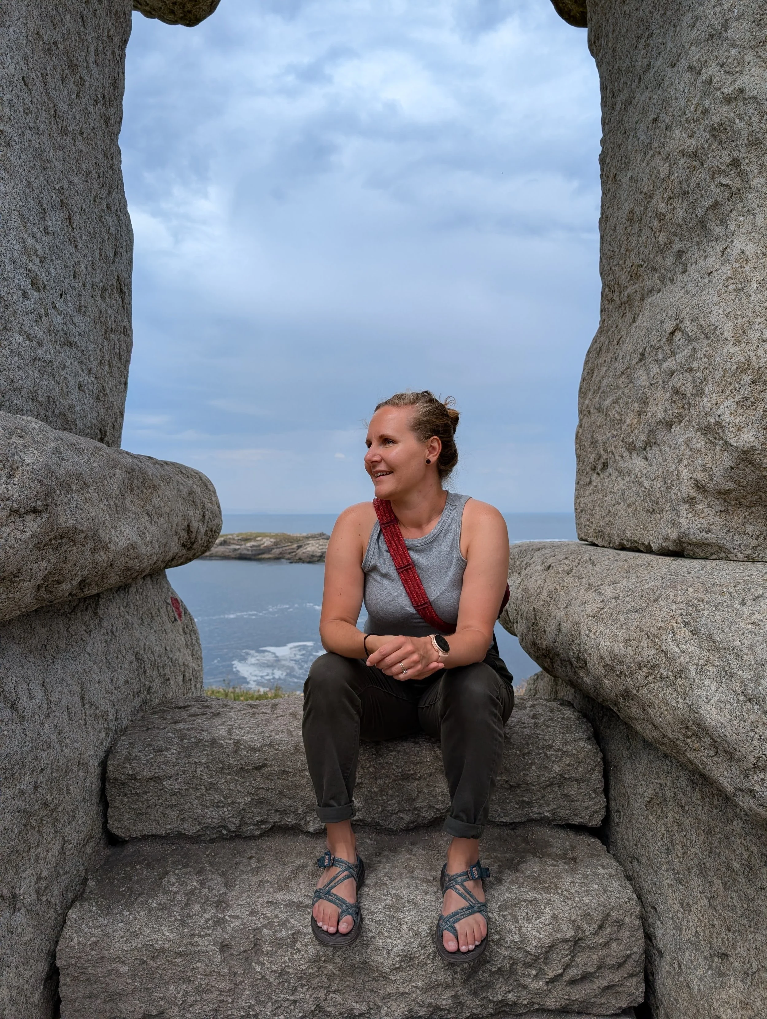 A woman sitting on a large stone structure, smiling, with the ocean and sky in the background.