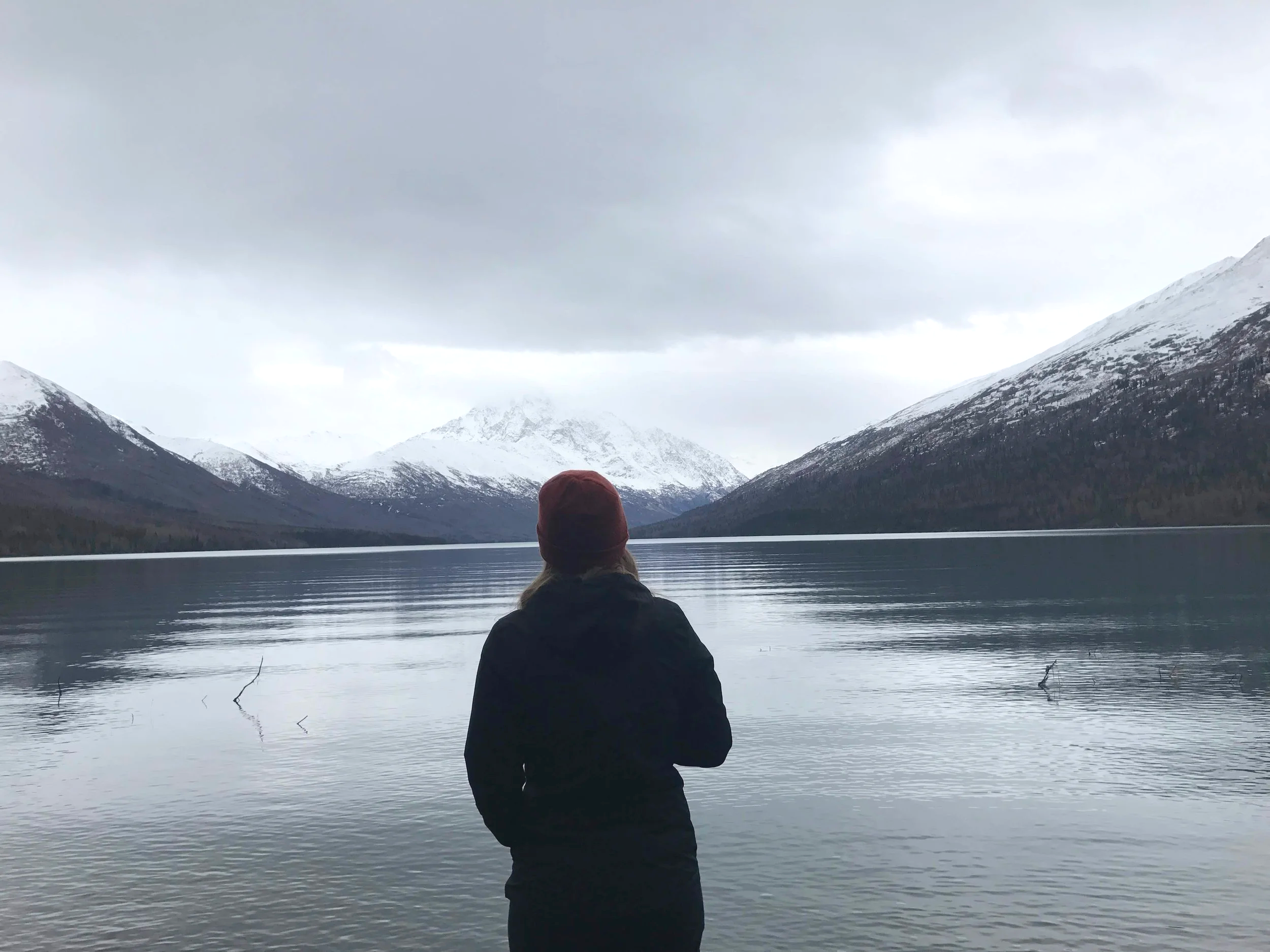Person with a red hat standing near a lake with snow-capped mountains in the background under a cloudy sky.