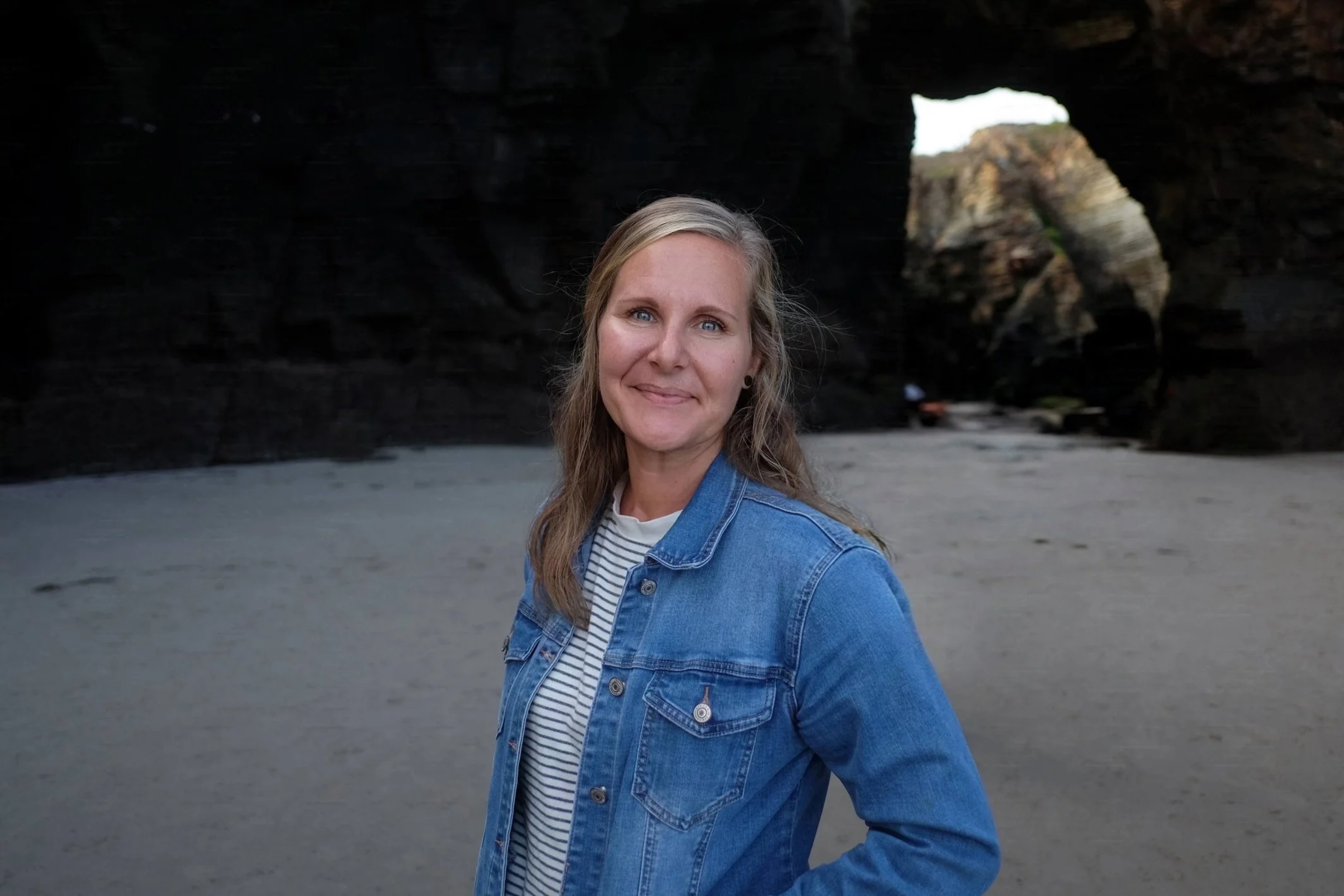 A woman with blonde hair and blue eyes standing on a beach with rocky formations and caves in the background.