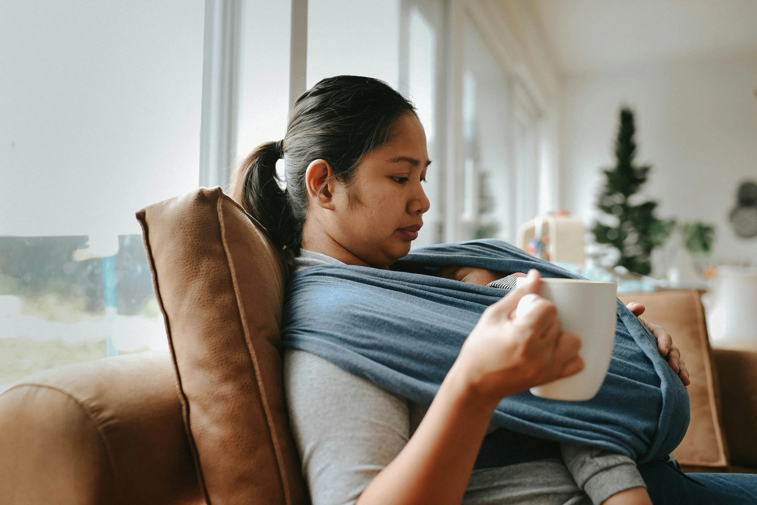 A woman sitting on a couch with a baby in a carrier strapped to her chest, holding a mug, in a room with a window and Christmas tree in the background.