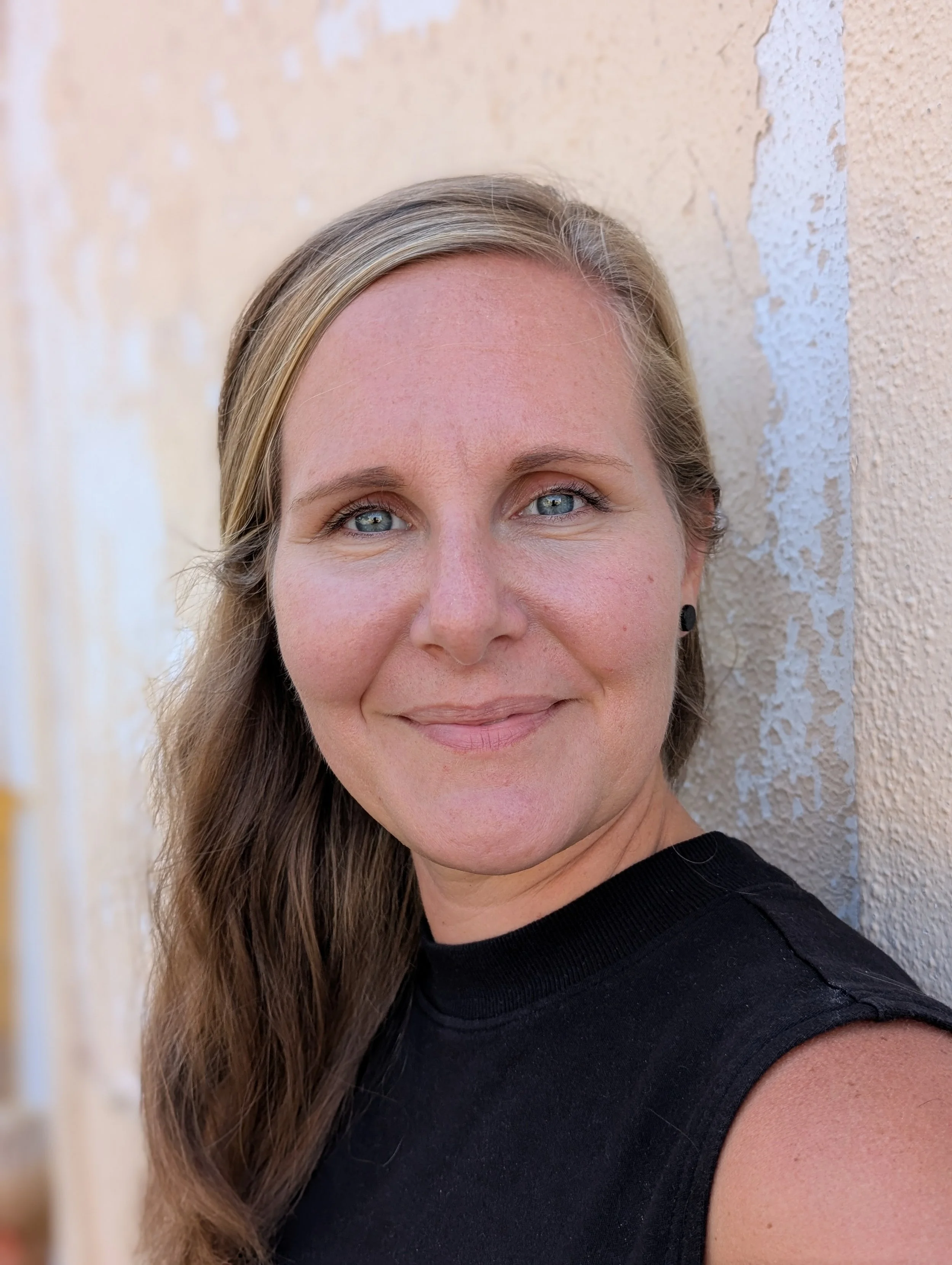 Close-up photo of a smiling woman with blonde hair and blue eyes, wearing a black sleeveless top, standing against a beige textured wall.
