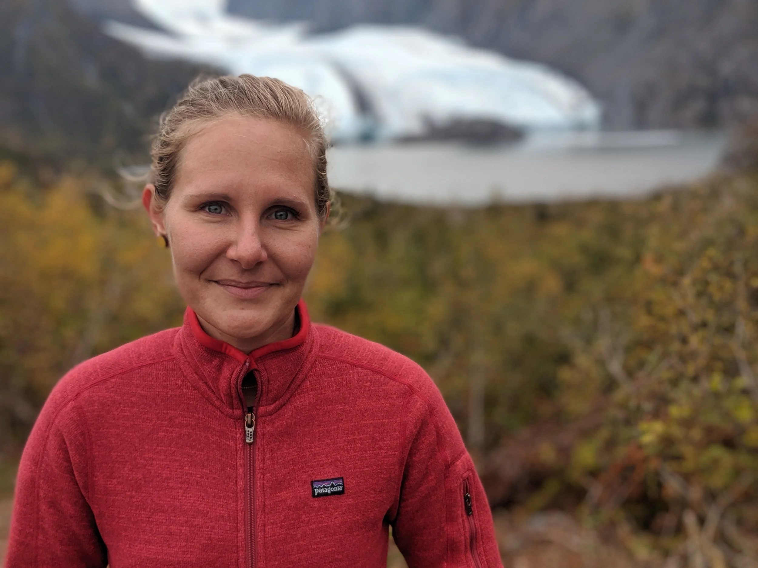 A woman in a red Patagonia jacket standing outdoors with autumn trees and a glacier in the background.