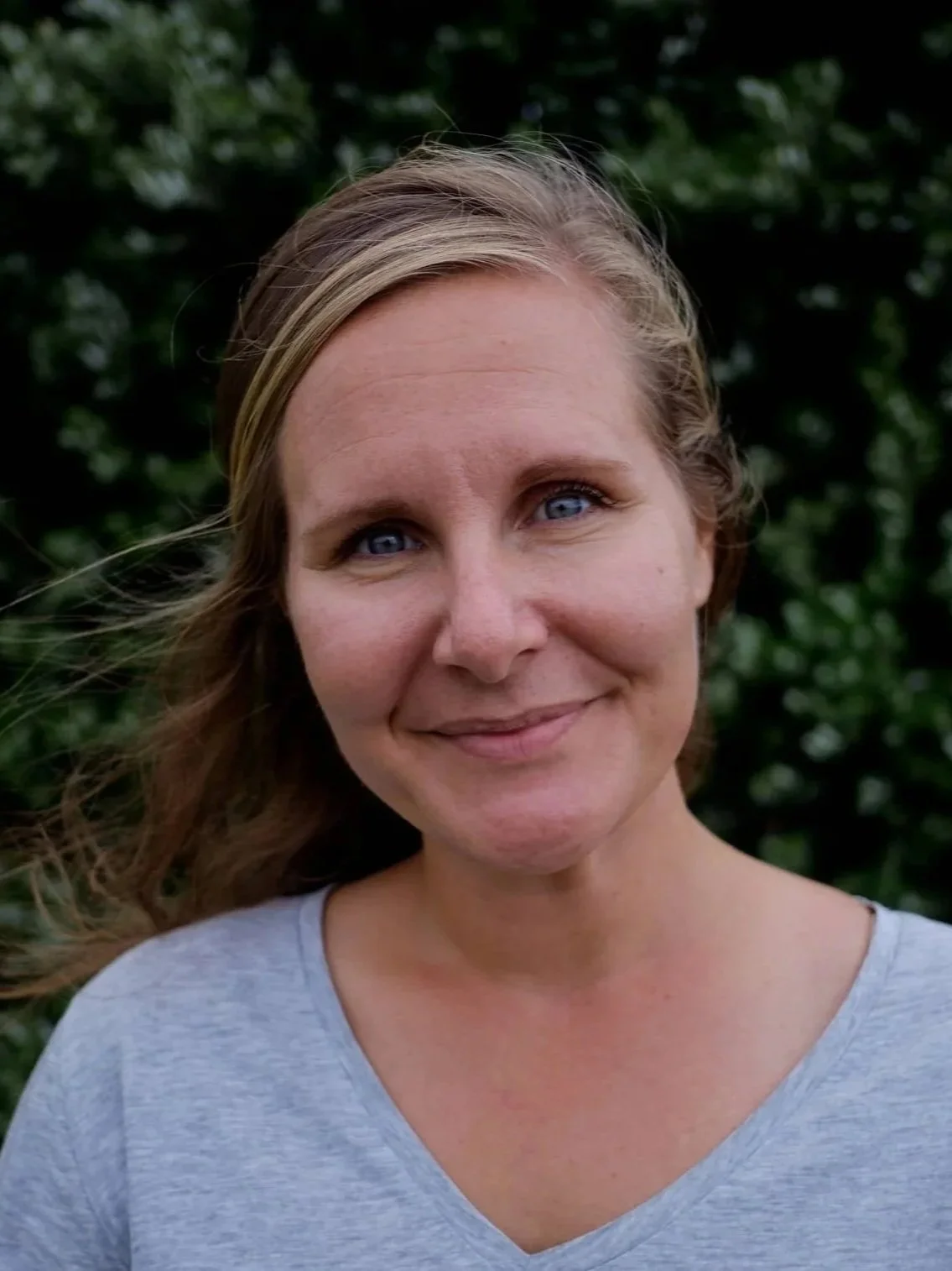 A woman with blue eyes and light brown hair standing outdoors with greenery in the background, wearing a gray shirt and smiling.