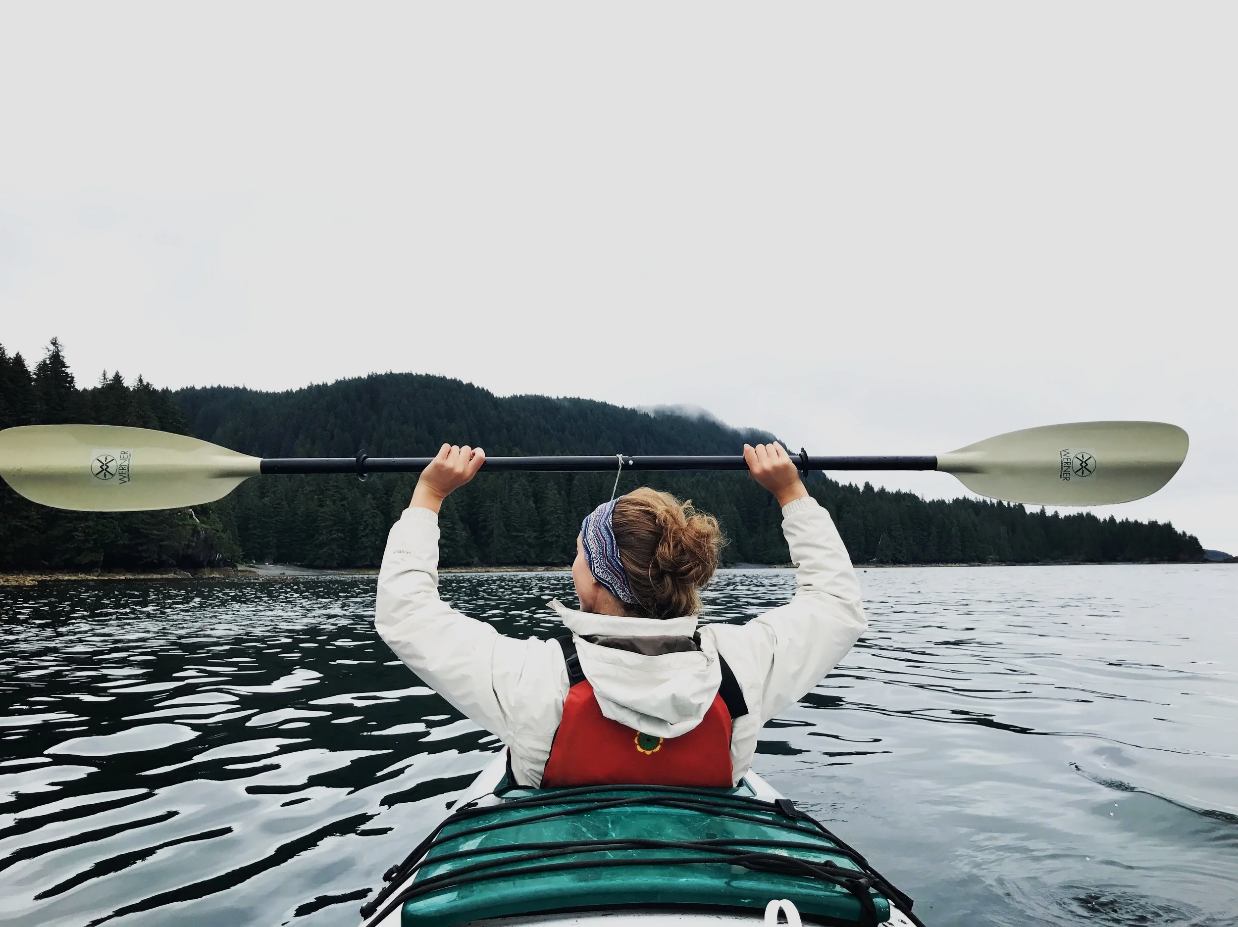 Person in a kayak holding a double-bladed paddle, with a forested mountain landscape in the background.