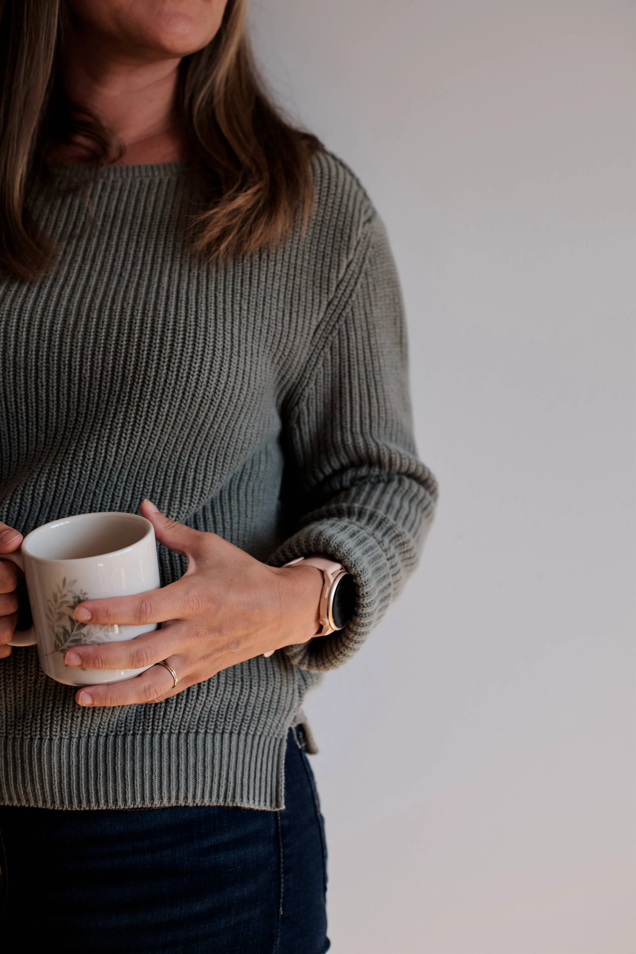 A woman wearing a gray knit sweater holding a mug with a floral design, standing against a plain wall.
