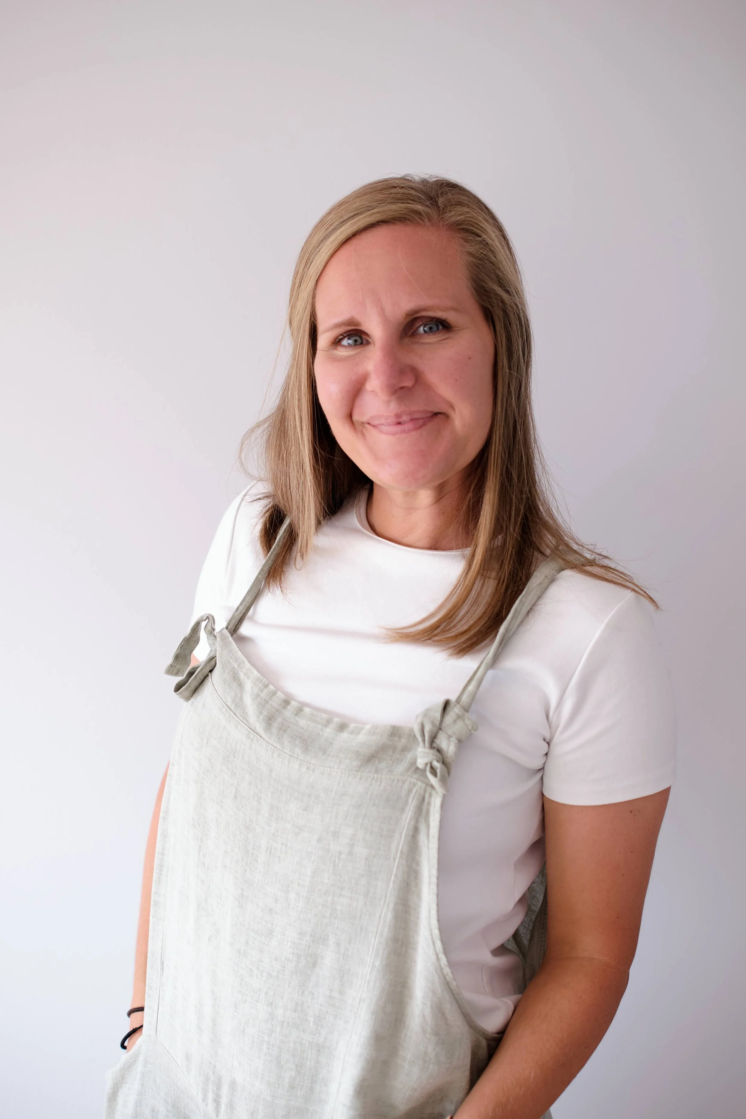A woman with light brown hair smiling wearing a white shirt and a light-colored apron, standing against a plain, light gray background.