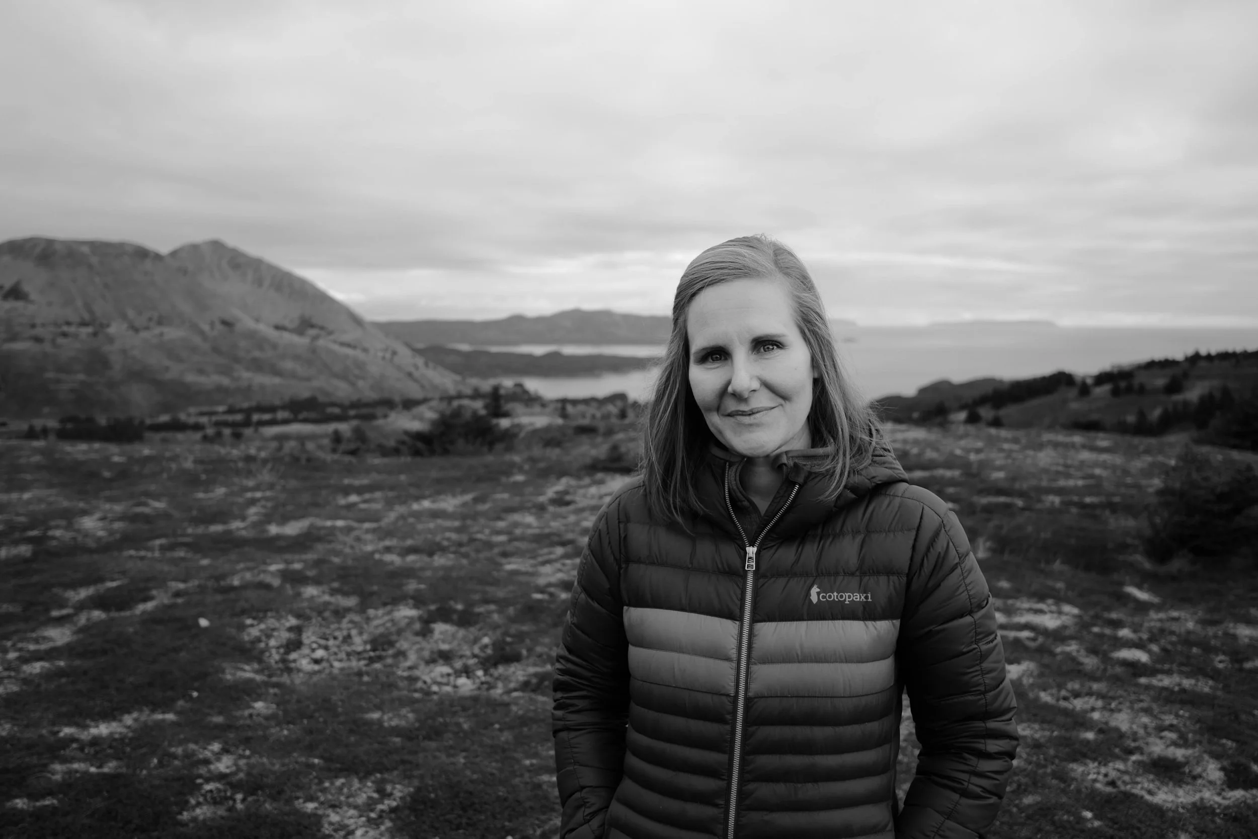 A woman with shoulder-length hair wearing a puffy jacket standing outdoors in a rugged landscape with mountains and a body of water in the background
