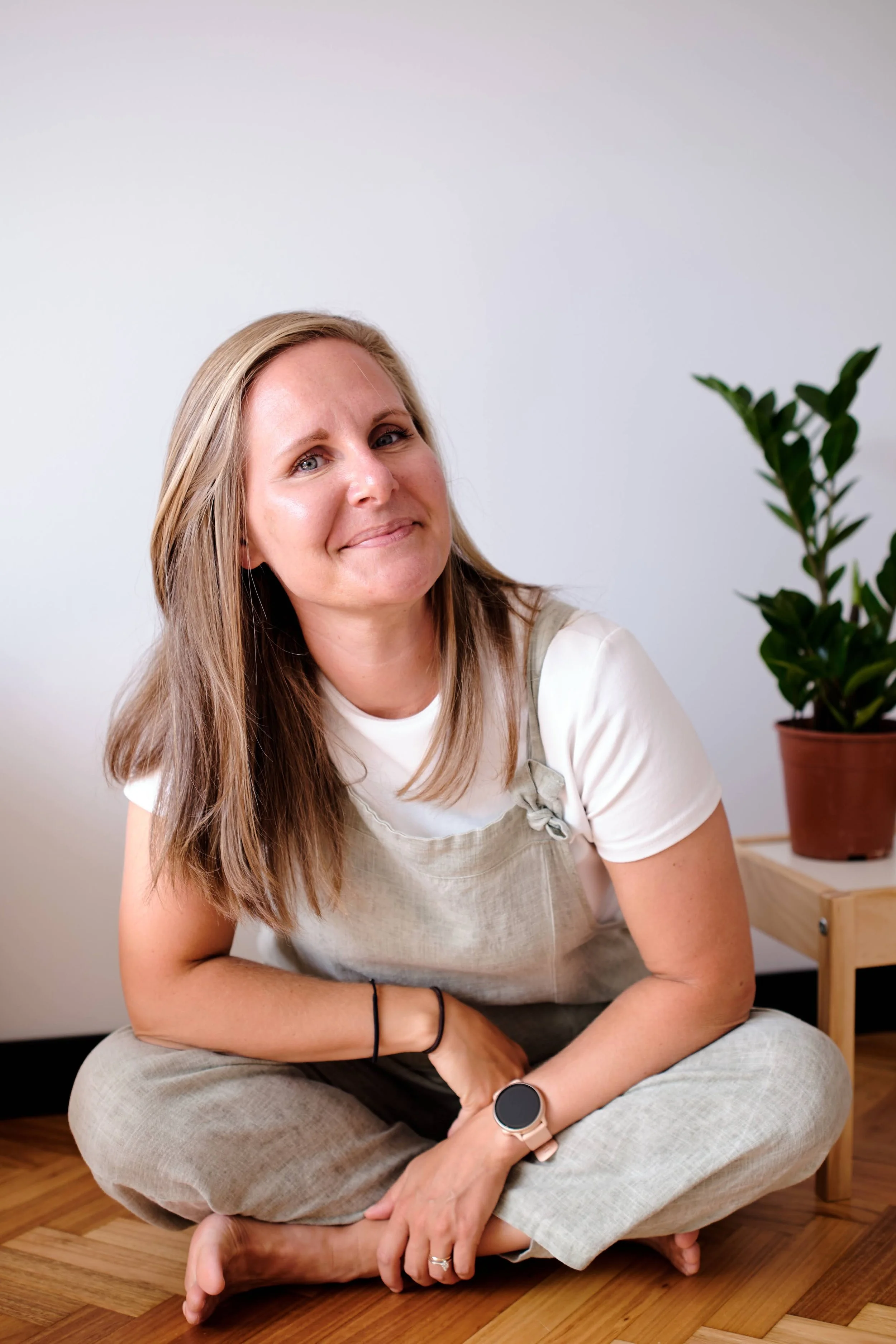 A woman with long light brown hair sitting cross-legged on the wooden floor, smiling at the camera. She is wearing a white t-shirt,overalls, and a smartwatch on her left wrist. There is a green leafy plant in a pot on a small table behind her to the right.