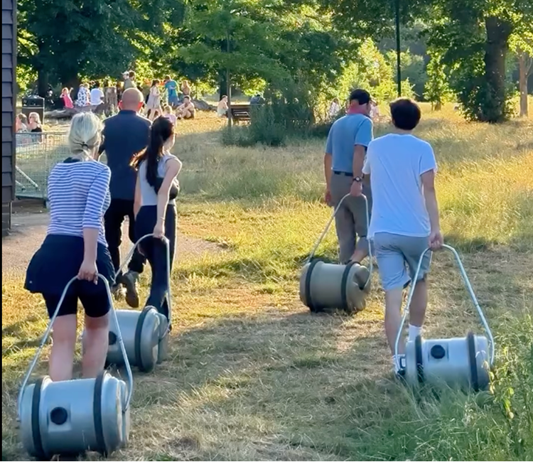 Four people, two women and two men, walking through a grassy park area, each pulling small cylindrical personal sound systems on wheels. There are other people and trees in the background, suggesting a park or outdoor event.