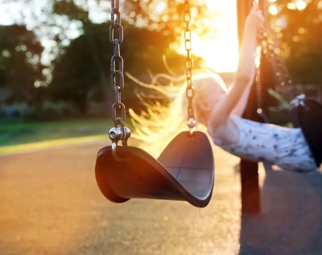 A close-up of a playground swing with a child swinging and the sun setting in the background.
