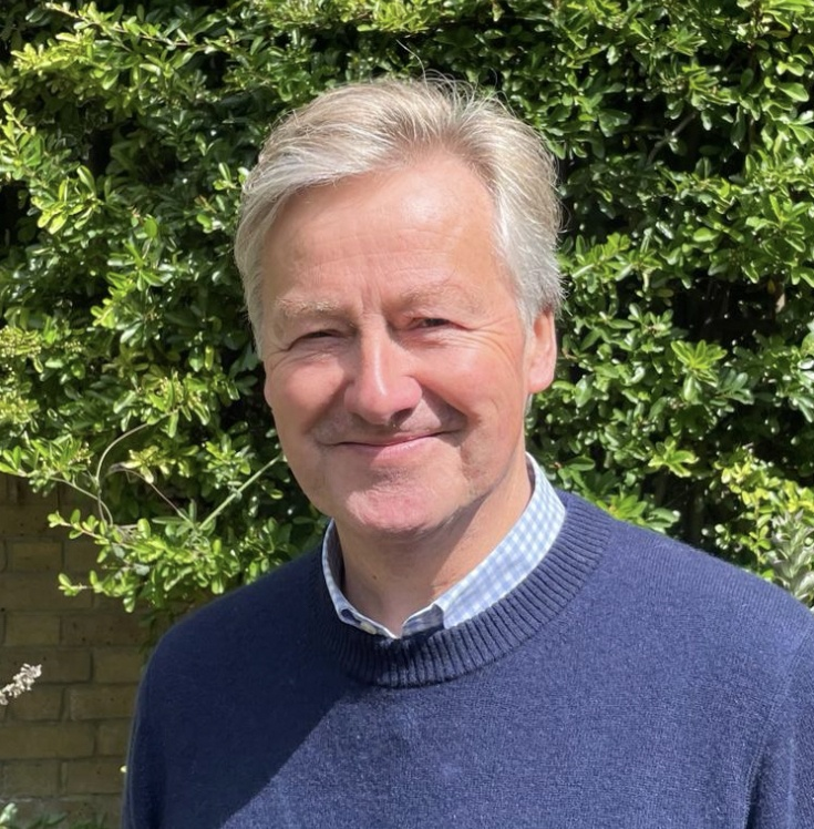A smiling middle-aged man with gray hair in a blue sweater and checkered collared shirt, standing outdoors in front of green bushes.