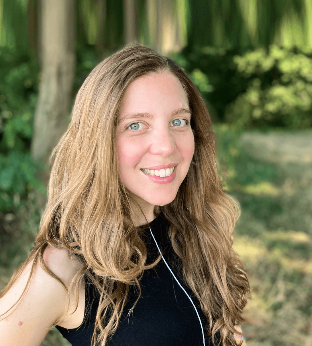 A young woman with long, wavy, light brown hair and blue eyes, smiling outdoors in a wooded area.