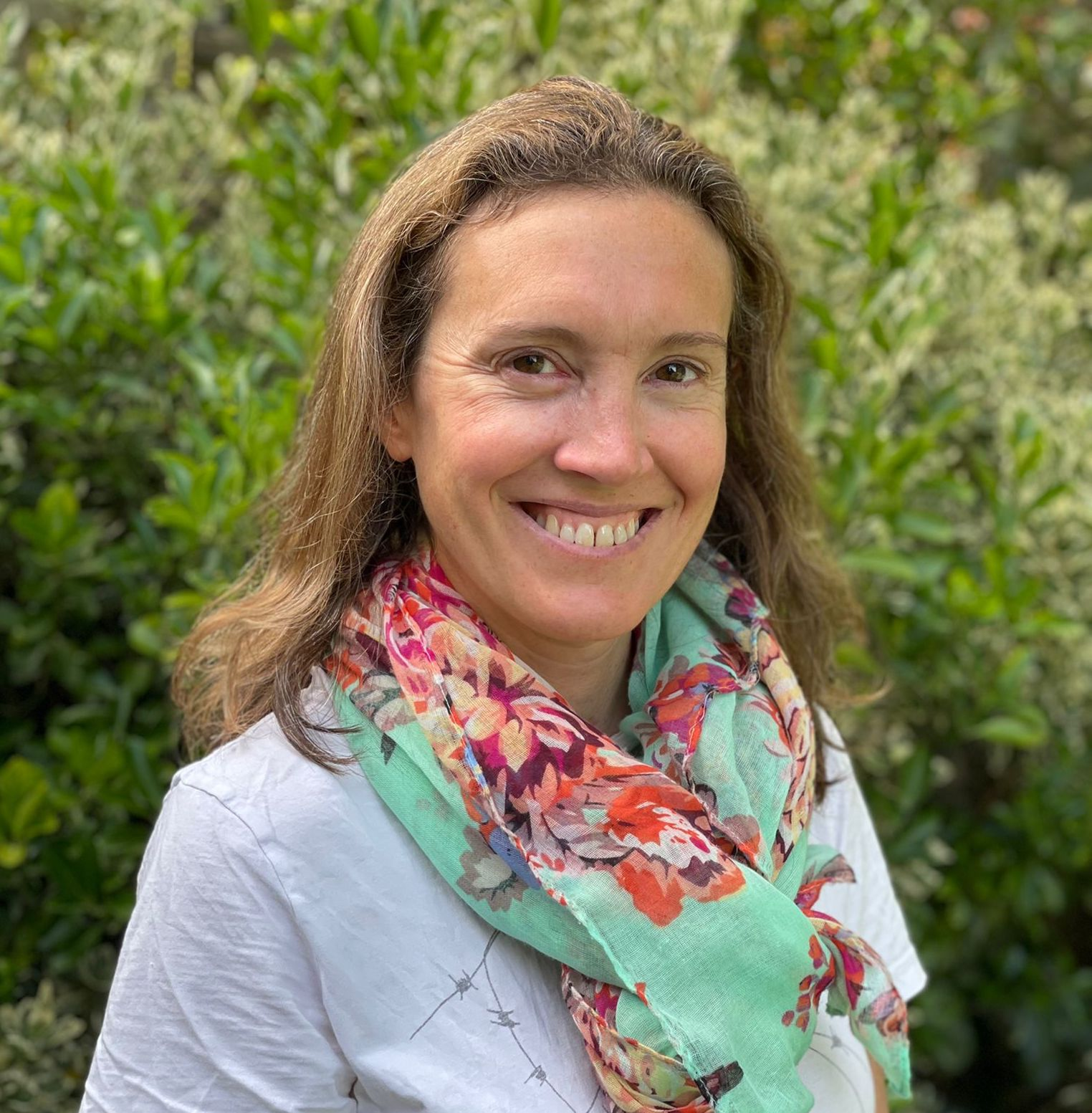 A woman with light brown hair smiling outdoors, wearing a white shirt and a colorful floral scarf, with green foliage in the background.