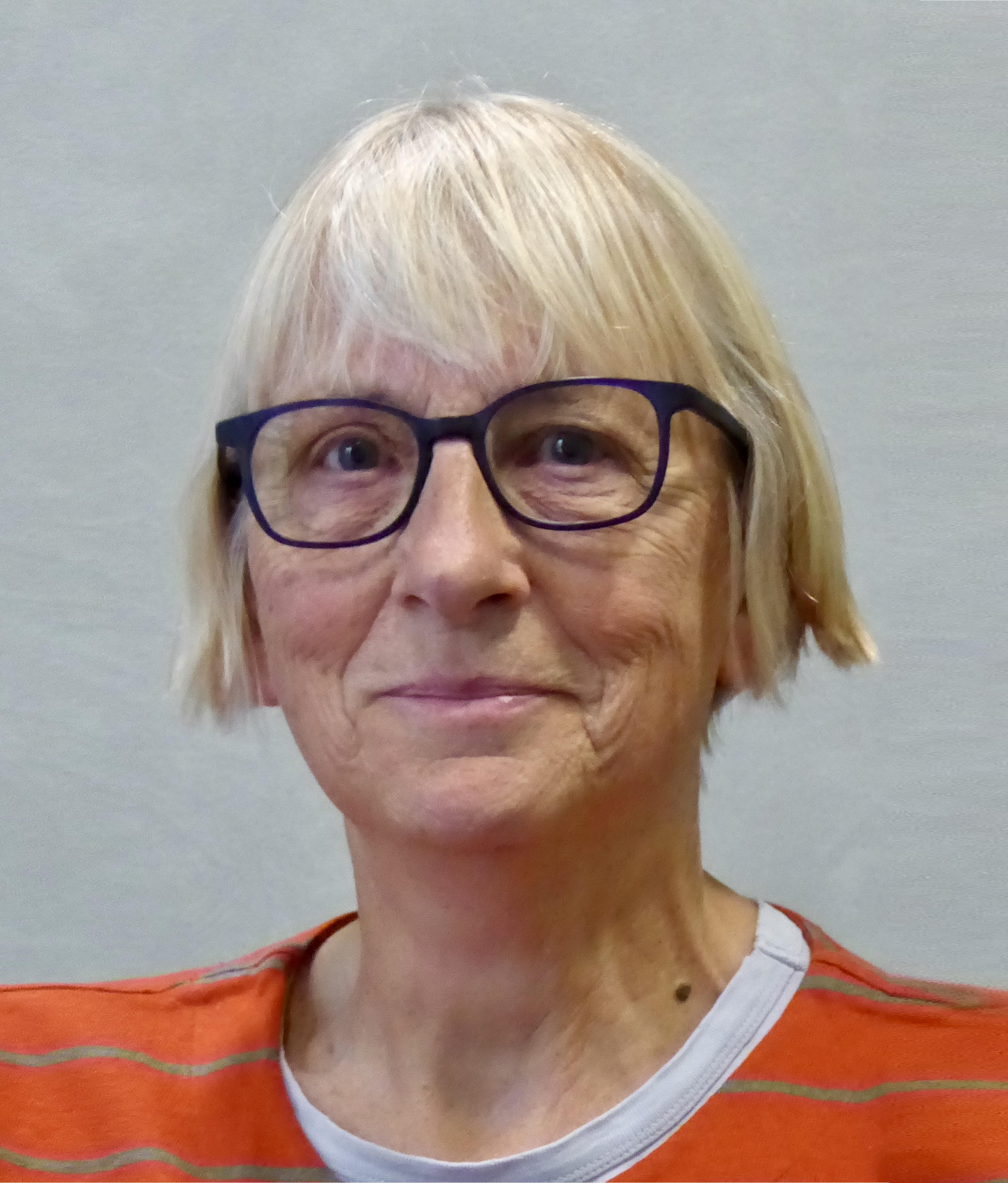 Close-up of a woman with short blond hair, wearing glasses and a red and white striped shirt, standing against a plain background.