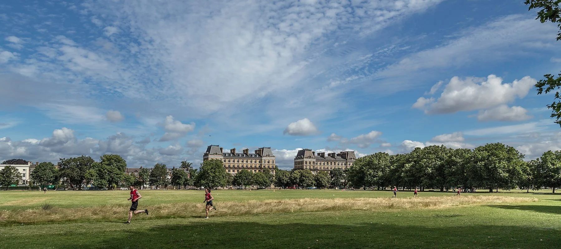 Park with green grass, trees, and buildings in the background under a partly cloudy sky. People are running and walking in the park.