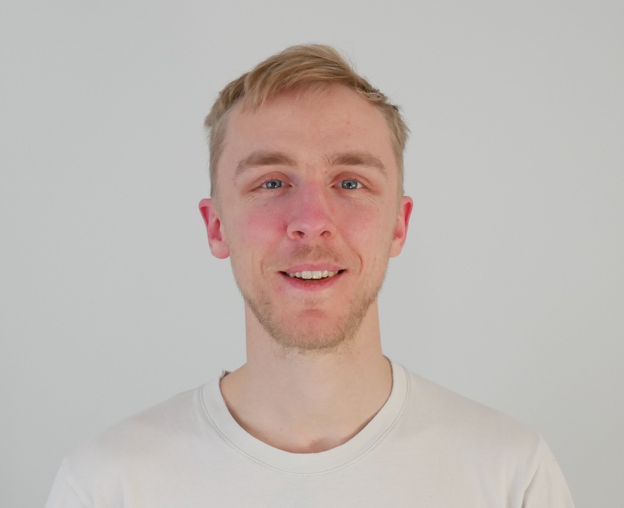 A young man with light blonde hair, blue eyes, and a light beard, smiling while wearing a white t-shirt, standing against a plain white background.