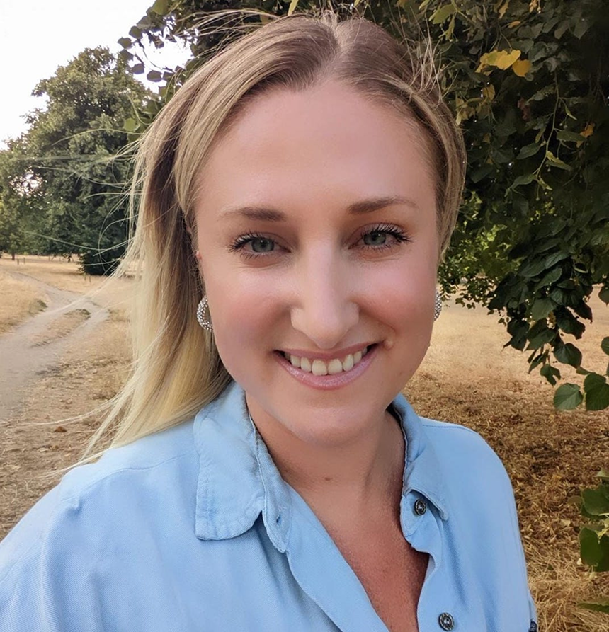 A young woman with long blond hair smiling at the camera outdoors, wearing a light blue shirt and round earrings, with trees and a dirt path in the background.